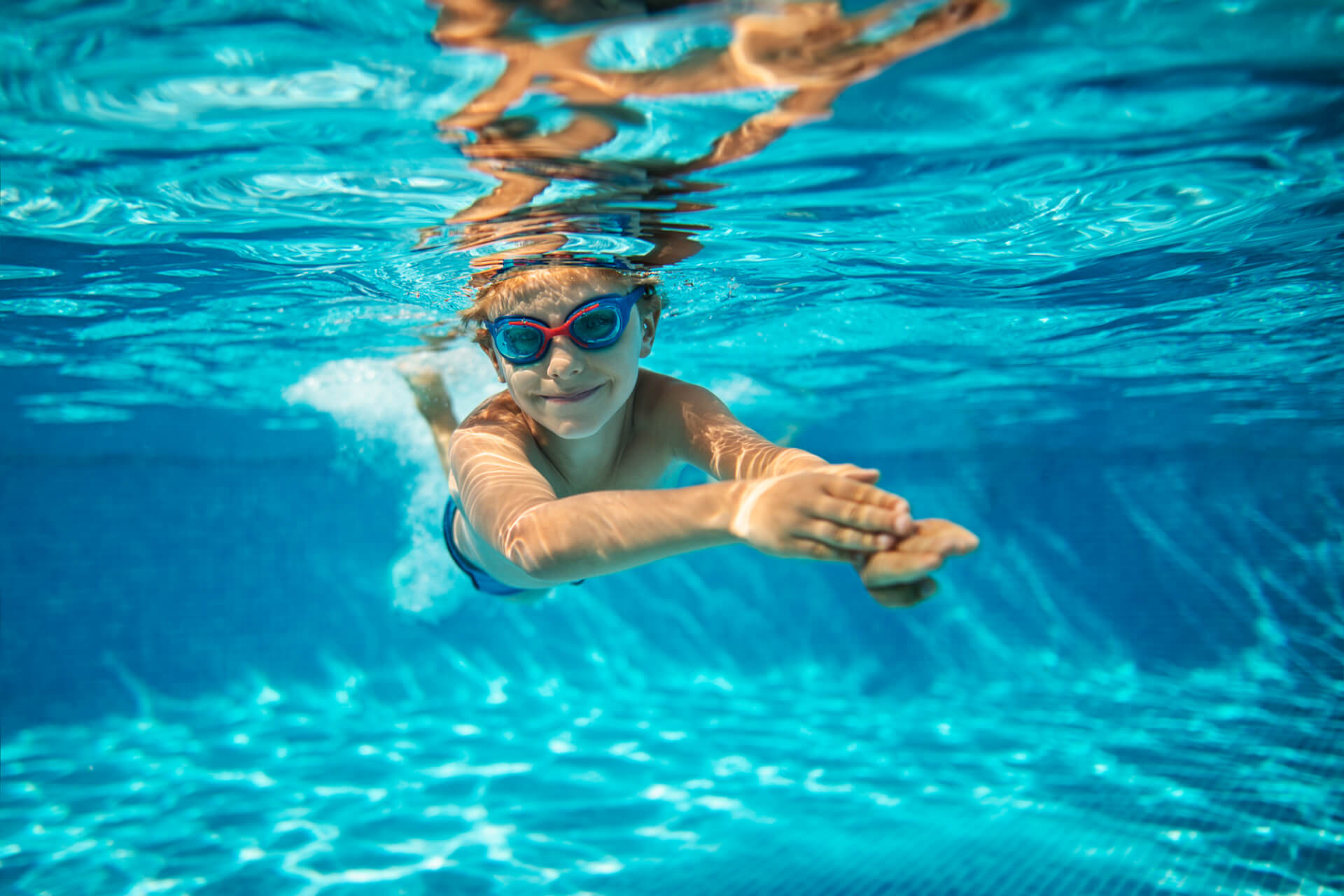 Un enfant portant des lunettes de natation nage sous l'eau dans une piscine, les bras tendus vers l'avant, la lumière du soleil créant des motifs sur l'eau bleue.