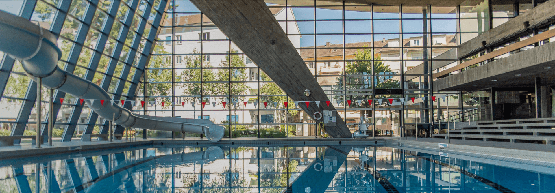 Hallenbad mit Wasserrutsche, großen Fenstern, dreieckigem Stützbalken und Reihen von rot-weißen Wimpeln, die über dem Wasser hängen.