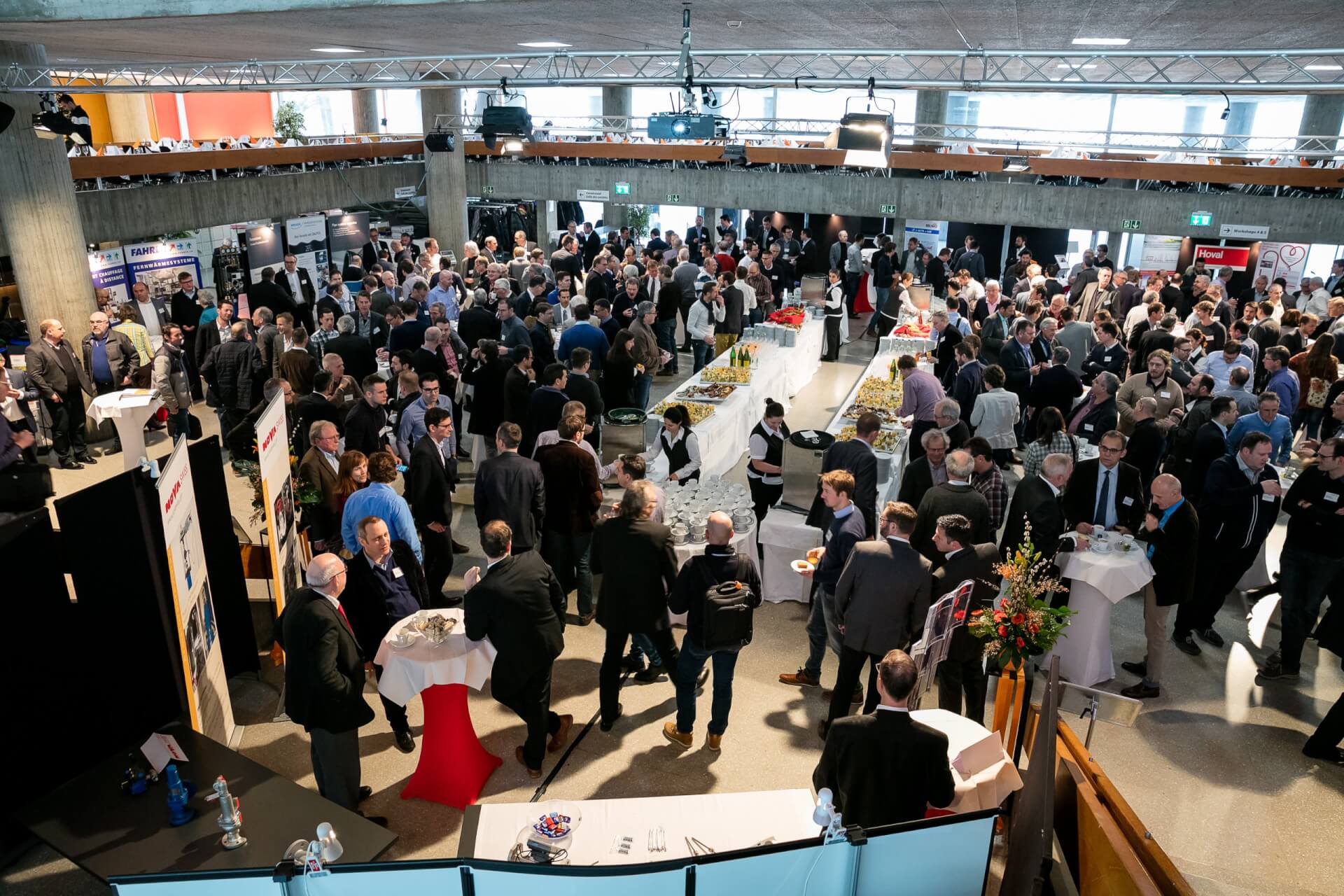 Eine große Gruppe von Menschen, die an einer Indoor-Networking-Veranstaltung teilnehmen, mit Tischen, Essen und Informationsdisplays, die überall im Raum zu sehen sind.