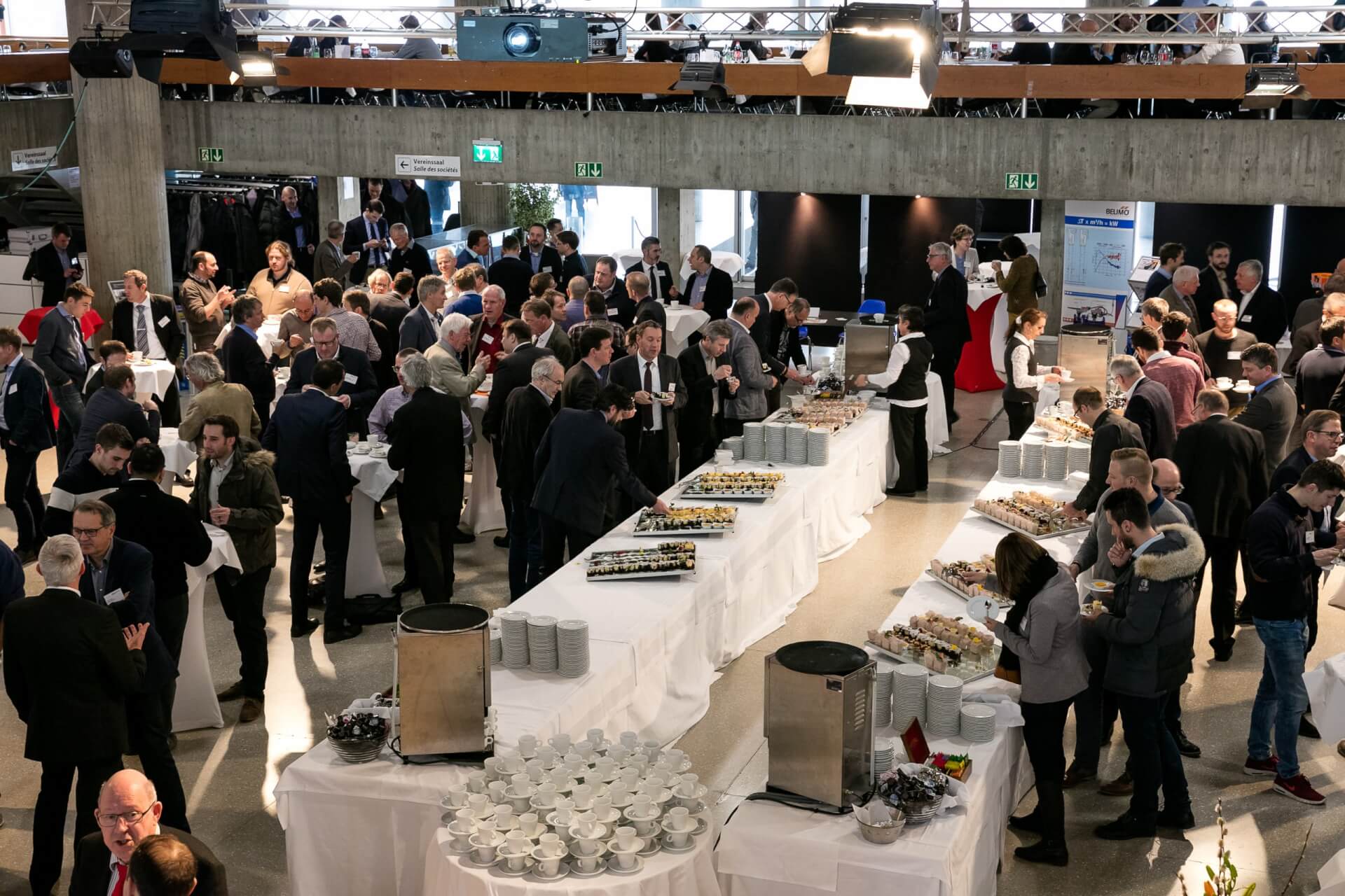 Eine große Gruppe von Menschen, die sich auf einer Konferenz oder einem Geschäftsevent austauschen, während in der Mitte des Raumes Buffet-Tische mit Speisen und Getränken aufgebaut sind.
