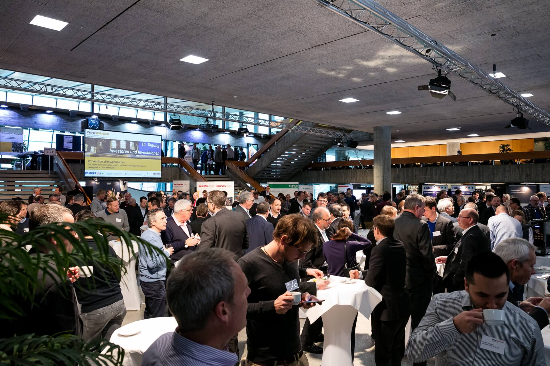 Eine große Gruppe von Personen nimmt an einer Networking-Veranstaltung oder Konferenz in einer modernen Halle mit Ständen, Tischen und Informationsdisplays teil.