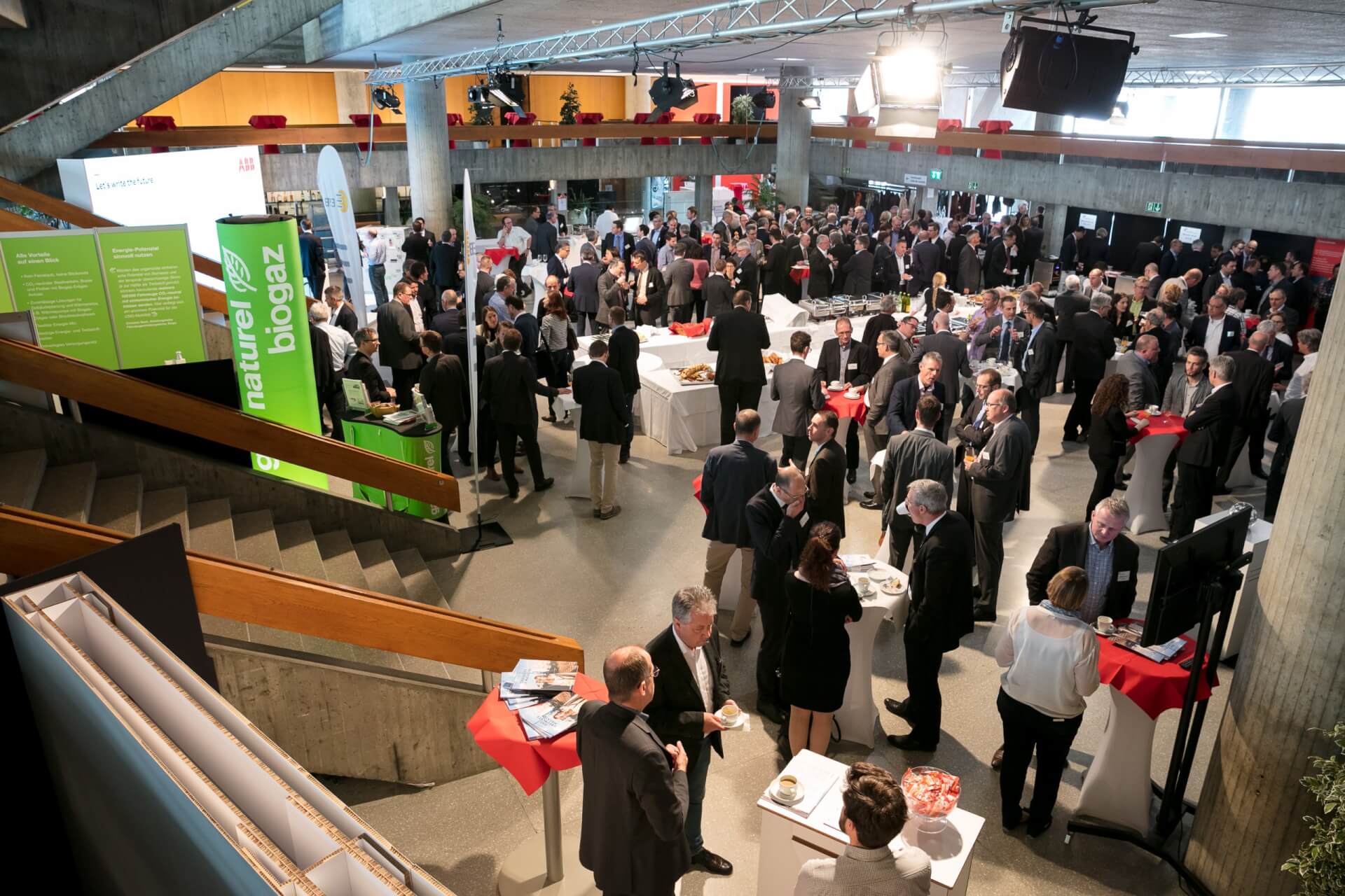 Eine große Gruppe von Personen in Geschäftskleidung nimmt an einer Networking-Veranstaltung in einem geräumigen, modernen Veranstaltungsort mit Informationsständen und Stehtischen teil.