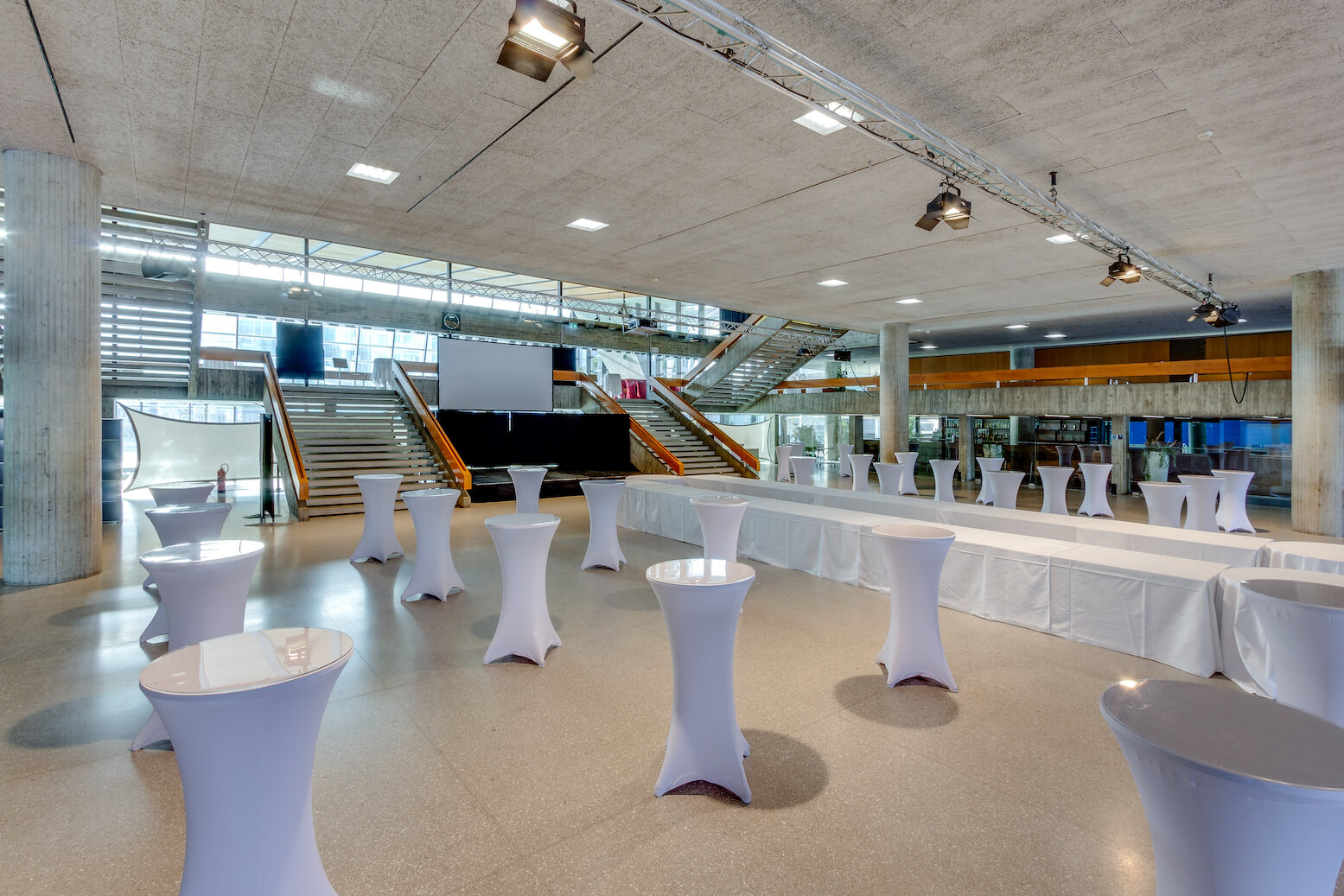 Moderner Veranstaltungssaal mit hohen Decken, überdachten Cocktailtischen, langen Buffettischen und einer großen Treppe, die zu einer Projektionsfläche im hinteren Bereich führt.