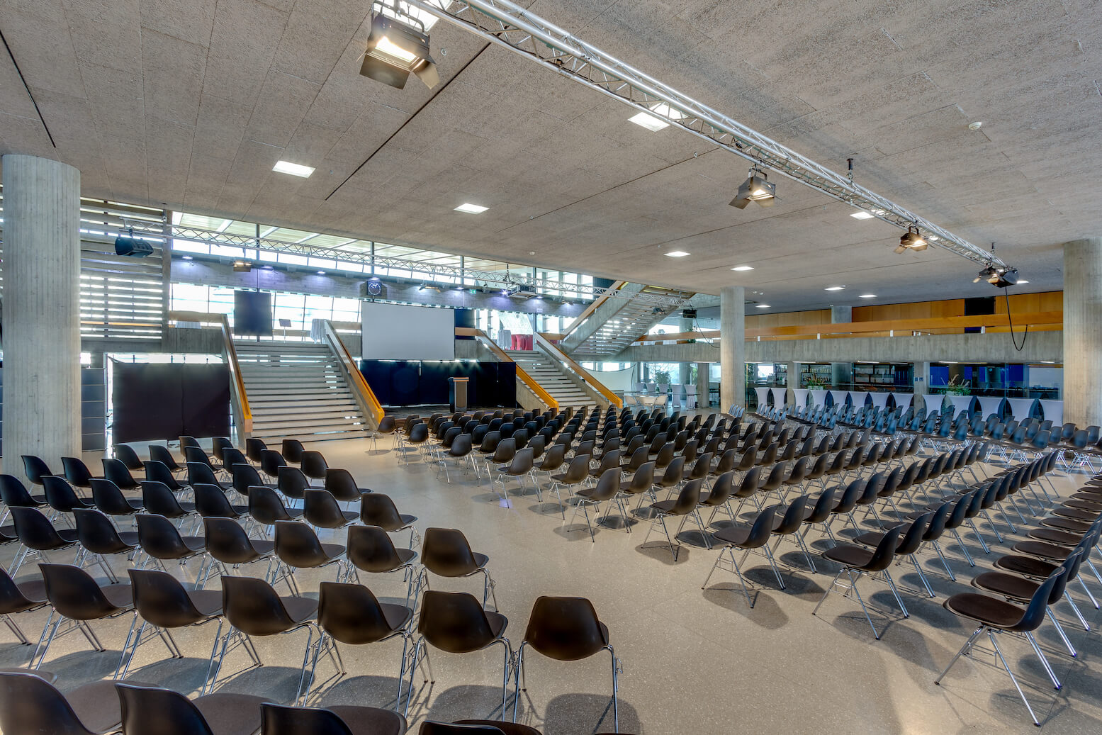 Großes modernes Auditorium mit Reihen leerer schwarzer Stühle vor einer Bühne, einem Podium und einer Projektionsfläche, mit Treppen und Glaswänden im Hintergrund.