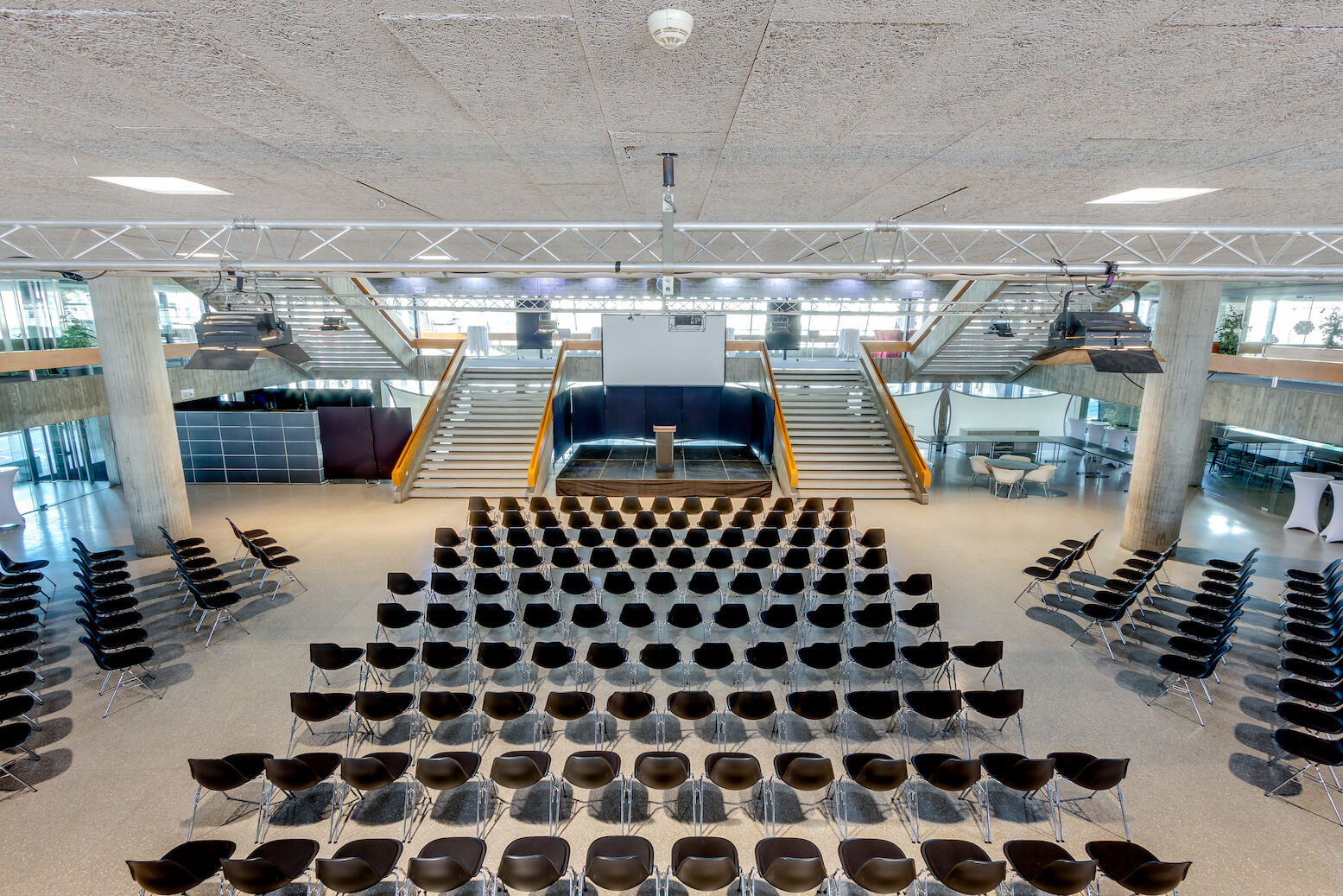 Reihen leerer schwarzer Stühle stehen vor einem Podium auf einer Bühne mit einer breiten Treppe und einem Glasgeländer im Hintergrund, in einem geräumigen, modernen Gebäude.