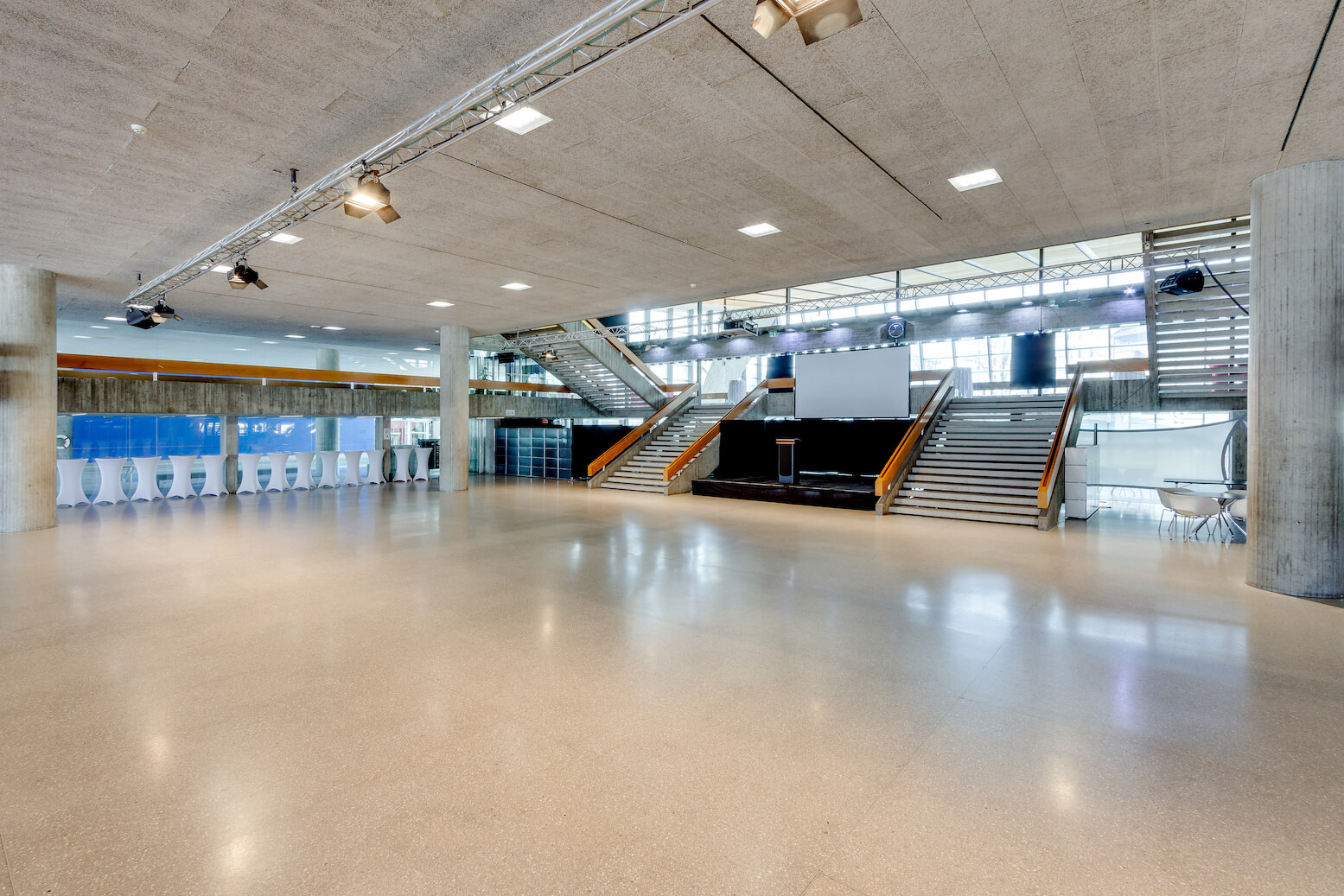 Geräumiges, modernes Foyer mit polierten Böden, zentraler Treppe, großer Projektionsfläche und hohen Fenstern, die viel Tageslicht hereinlassen. An einer Wand auf der linken Seite sind Urinale zu sehen.