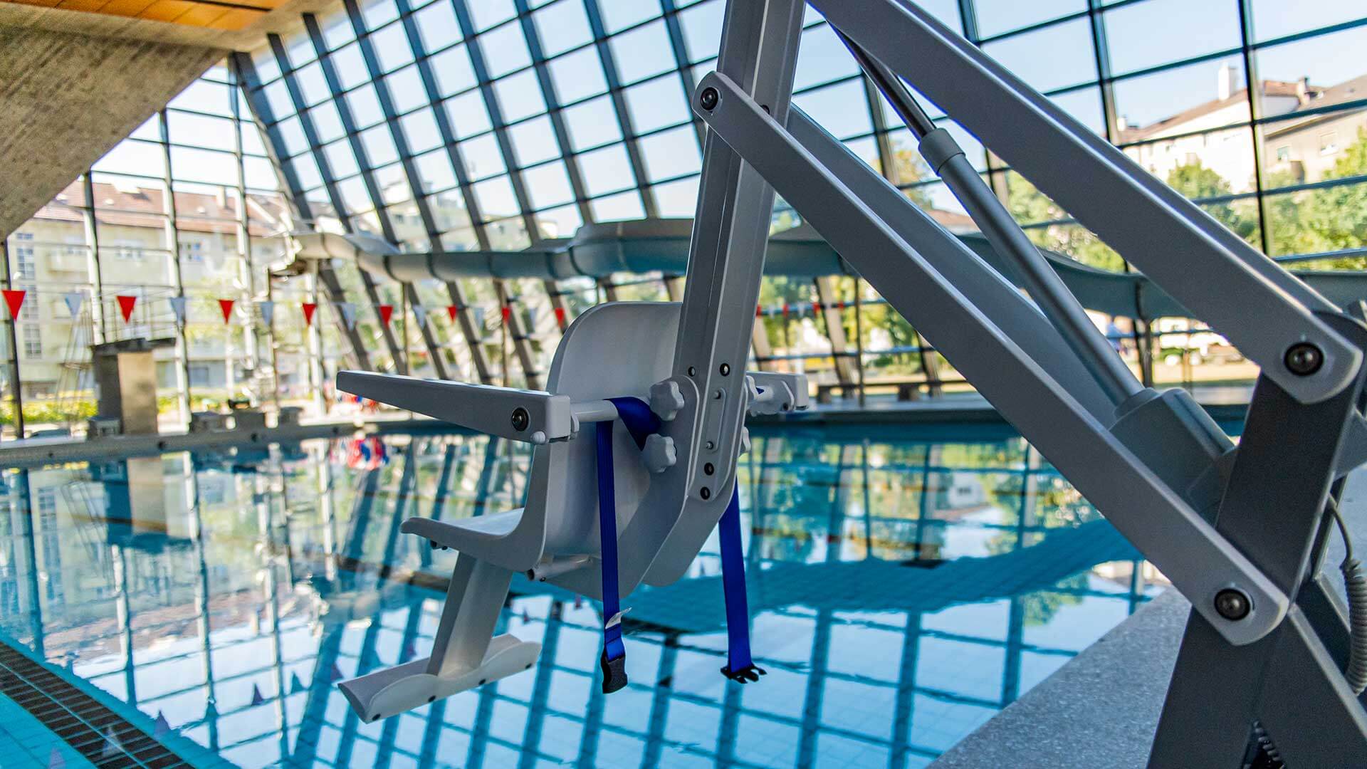 Ein Pool-Lift-Sessel steht am Rande eines Hallenbads mit großen Fenstern im Hintergrund.