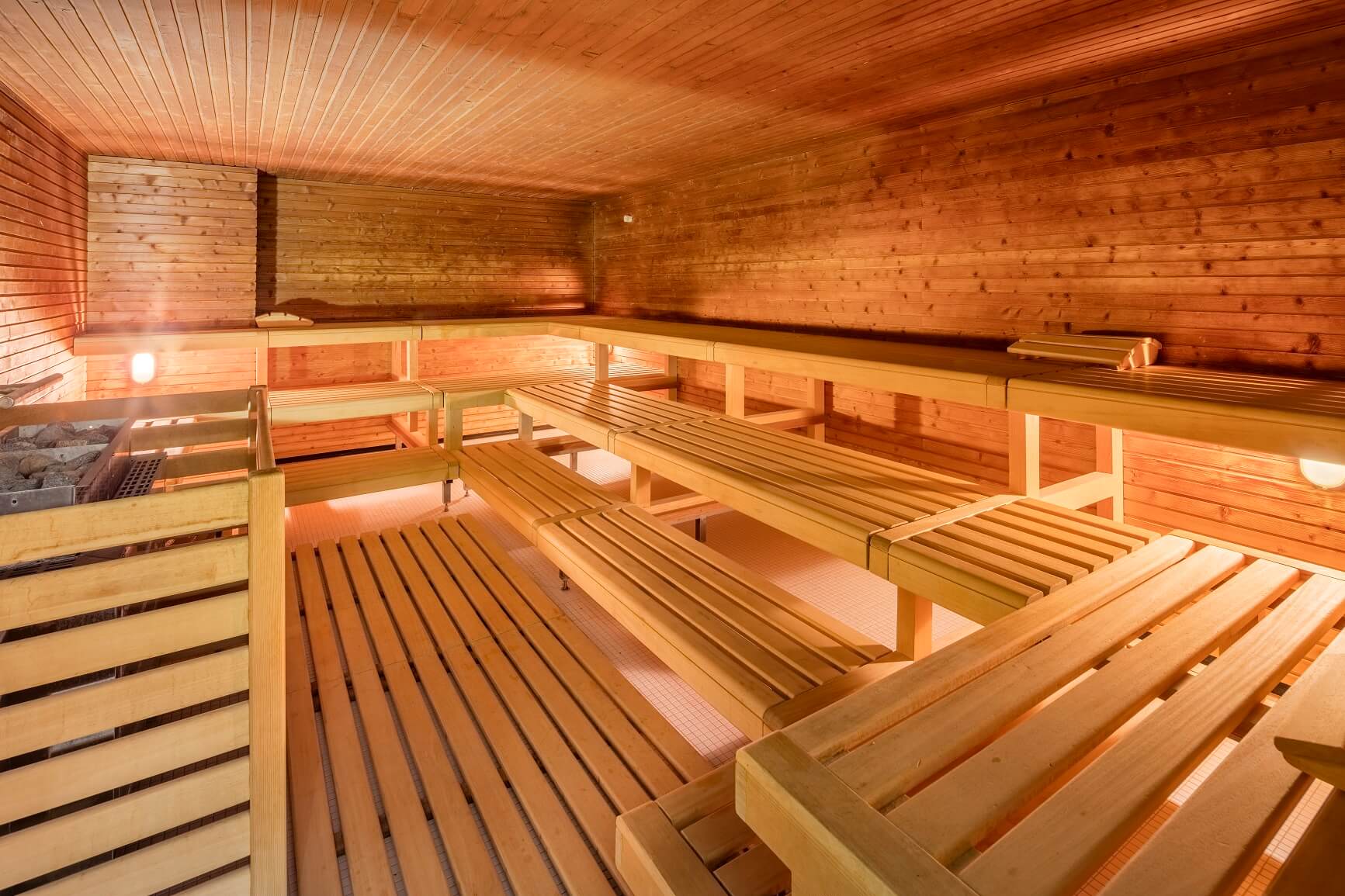 Großer hölzerner Saunaraum mit abgestuften Bänken, gedämpfter Beleuchtung und einem Steinofen auf der linken Seite. Die Wände, die Decke und der Boden sind aus hellem Holz gefertigt.