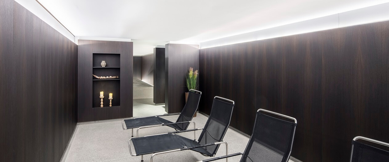 Minimalistischer Ruheraum mit dunkler Holzvertäfelung, drei schwarzen Loungesesseln, dekorativen Regalen und einer Pflanze in der Ecke.