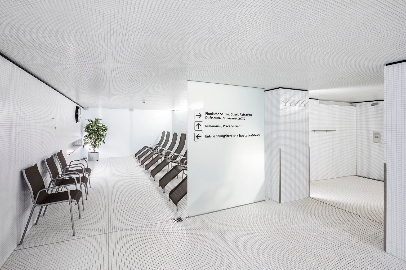 Ein moderner Spa-Entspannungsbereich mit Reihen von braunen Liegestühlen, weiß gefliesten Wänden und Böden, einer Topfpflanze und Wegweisern an der Wand.