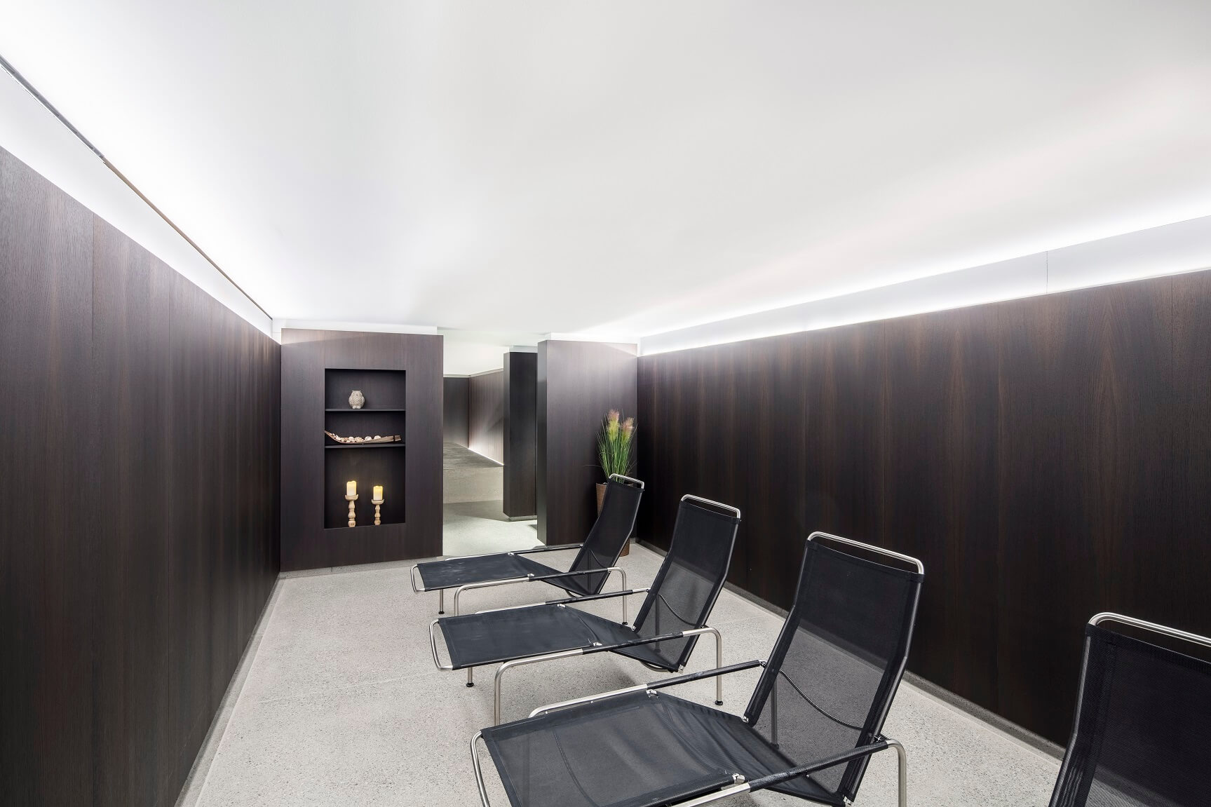 Ein minimalistischer Raum mit dunklen holzgetäfelten Wänden, drei schwarzen Lehnstühlen, einem kleinen Regal mit Kerzen und sanfter Deckenbeleuchtung.