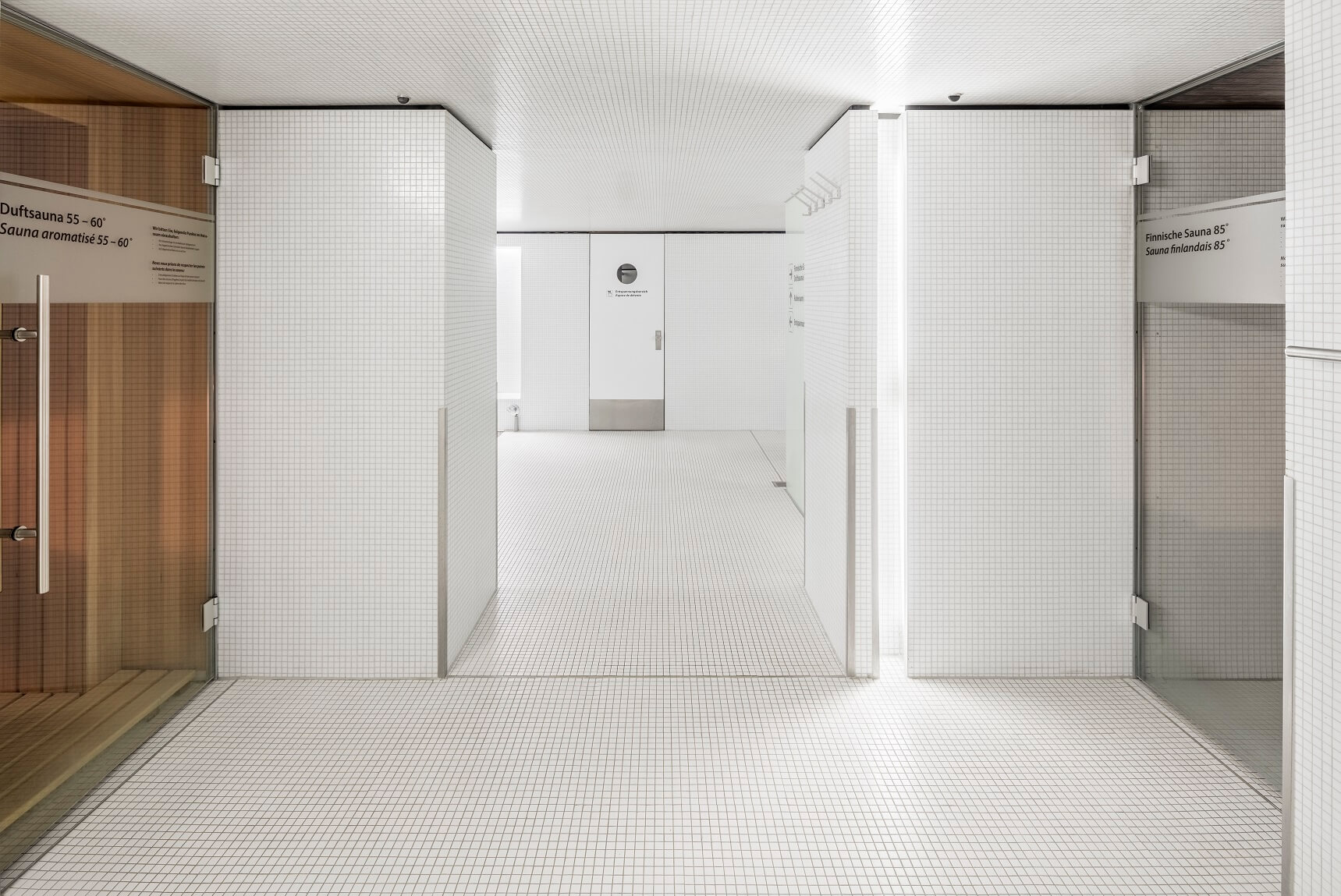 Minimalistischer, weiß gefliester Spa- oder Wellnessbereich mit Glastüren, die zu Saunaräumen auf jeder Seite und einem Flur in der Mitte führen.
