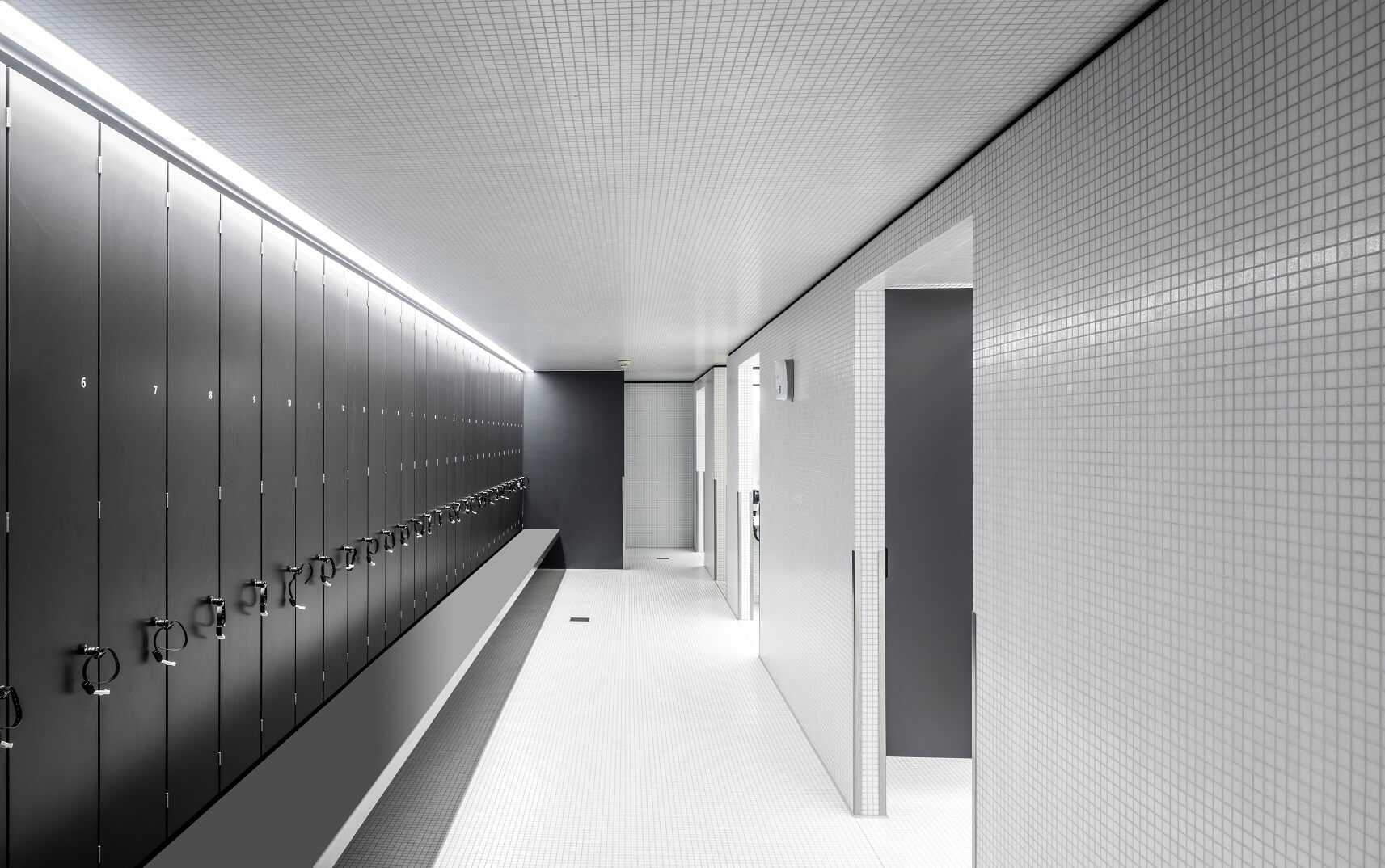 Ein moderner Umkleideraum mit dunkelgrauen Spinden auf der linken Seite, einem weiß gefliesten Boden und weißen Wänden sowie einem minimalistischen Design mit heller Deckenbeleuchtung.