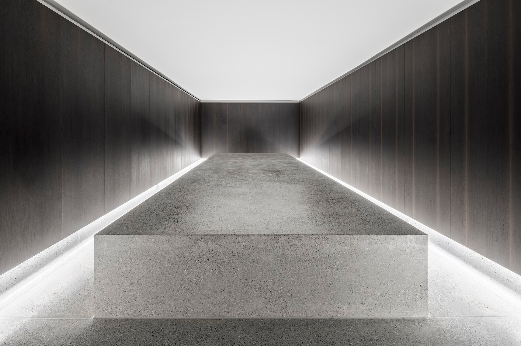 Ein minimalistischer Raum mit dunklen holzgetäfelten Wänden und einer großen, rechteckigen Betonplatte in der Mitte, die gleichmäßig und indirekt beleuchtet wird.