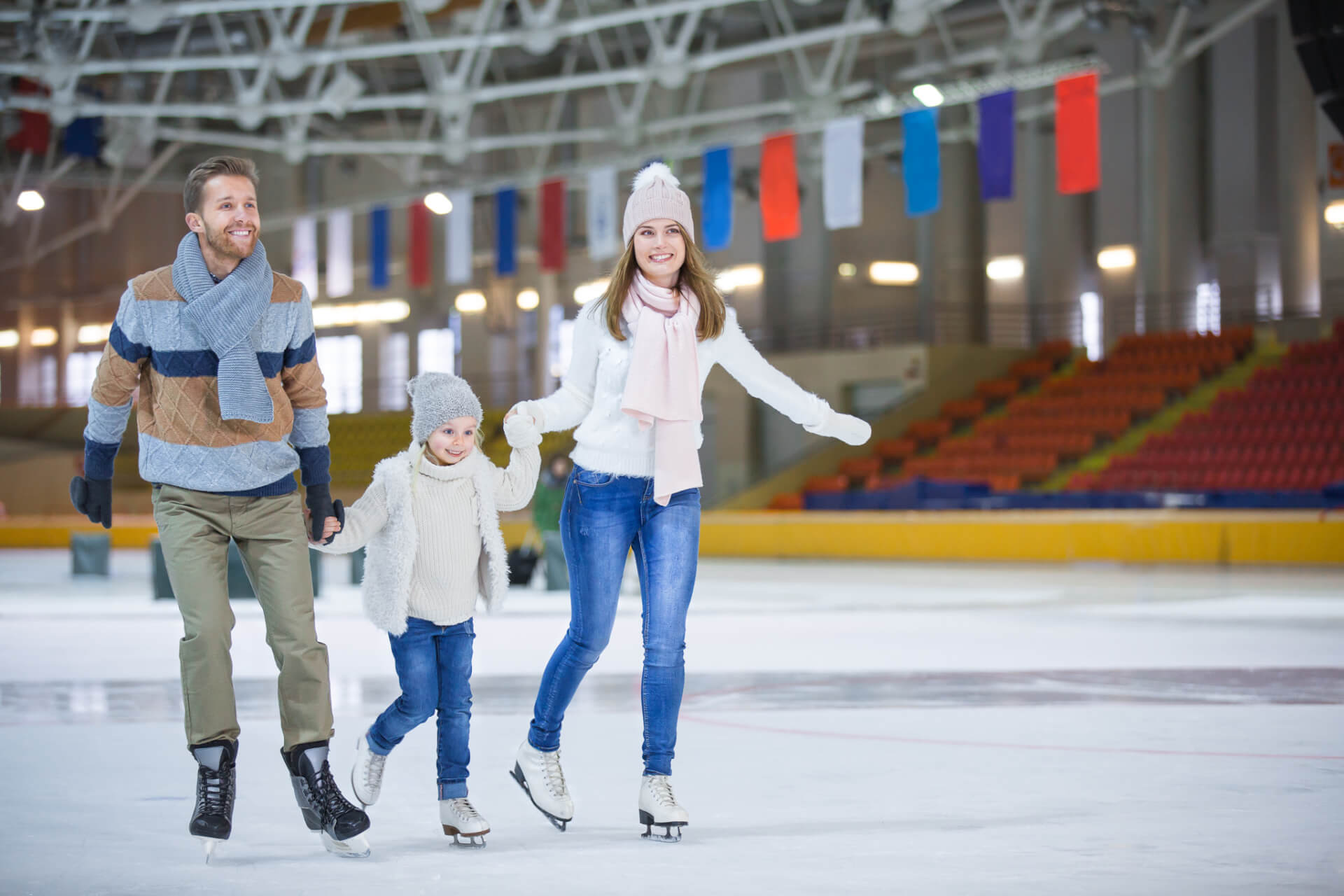 Eine dreiköpfige Familie in Winterkleidung läuft gemeinsam Schlittschuh auf einer Eisbahn mit leeren Stadionsitzen und hängenden Fahnen im Hintergrund.