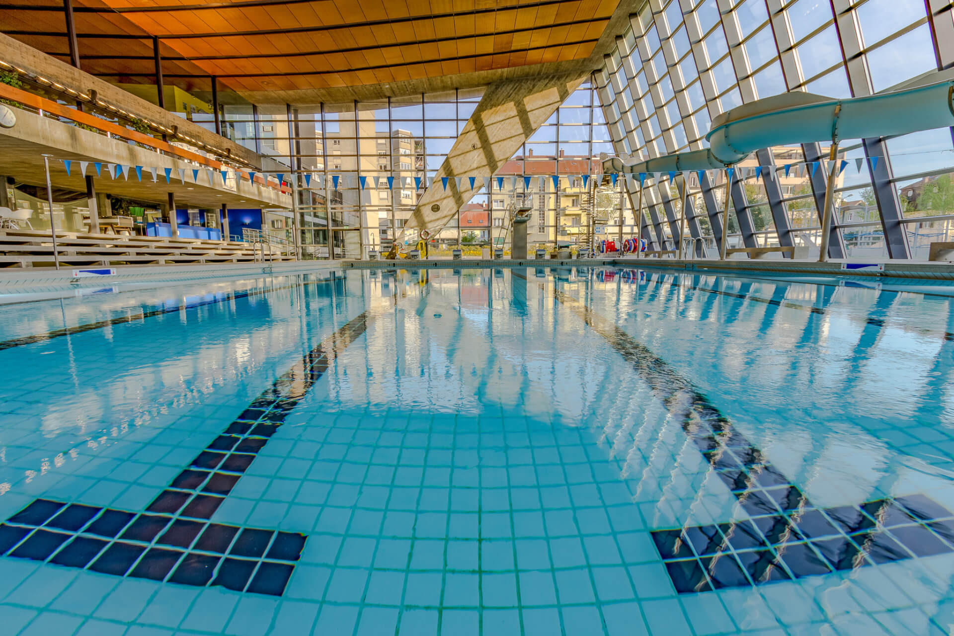 Hallenbad mit klarem Wasser, markierten Bahnen, einer Wasserrutsche, dreieckigen Fahnen und großen Glasfenstern, die Tageslicht hereinlassen. Durch die Fenster kann man die Gebäude draußen sehen.