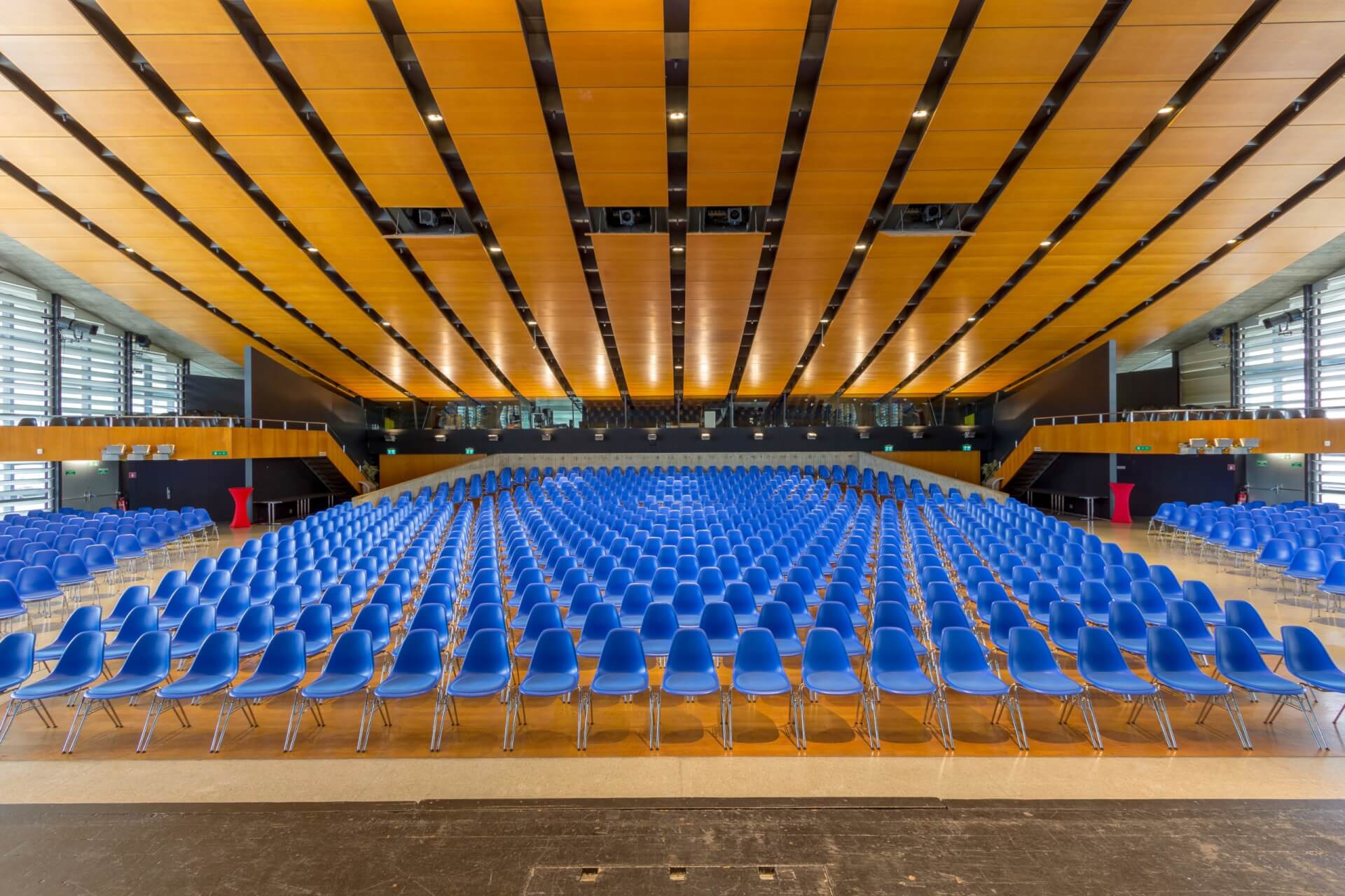 Großes Auditorium mit Reihen leerer blauer Stühle vor einer Bühne, hölzernen Deckenpaneelen und Balkonbestuhlung im Hintergrund.