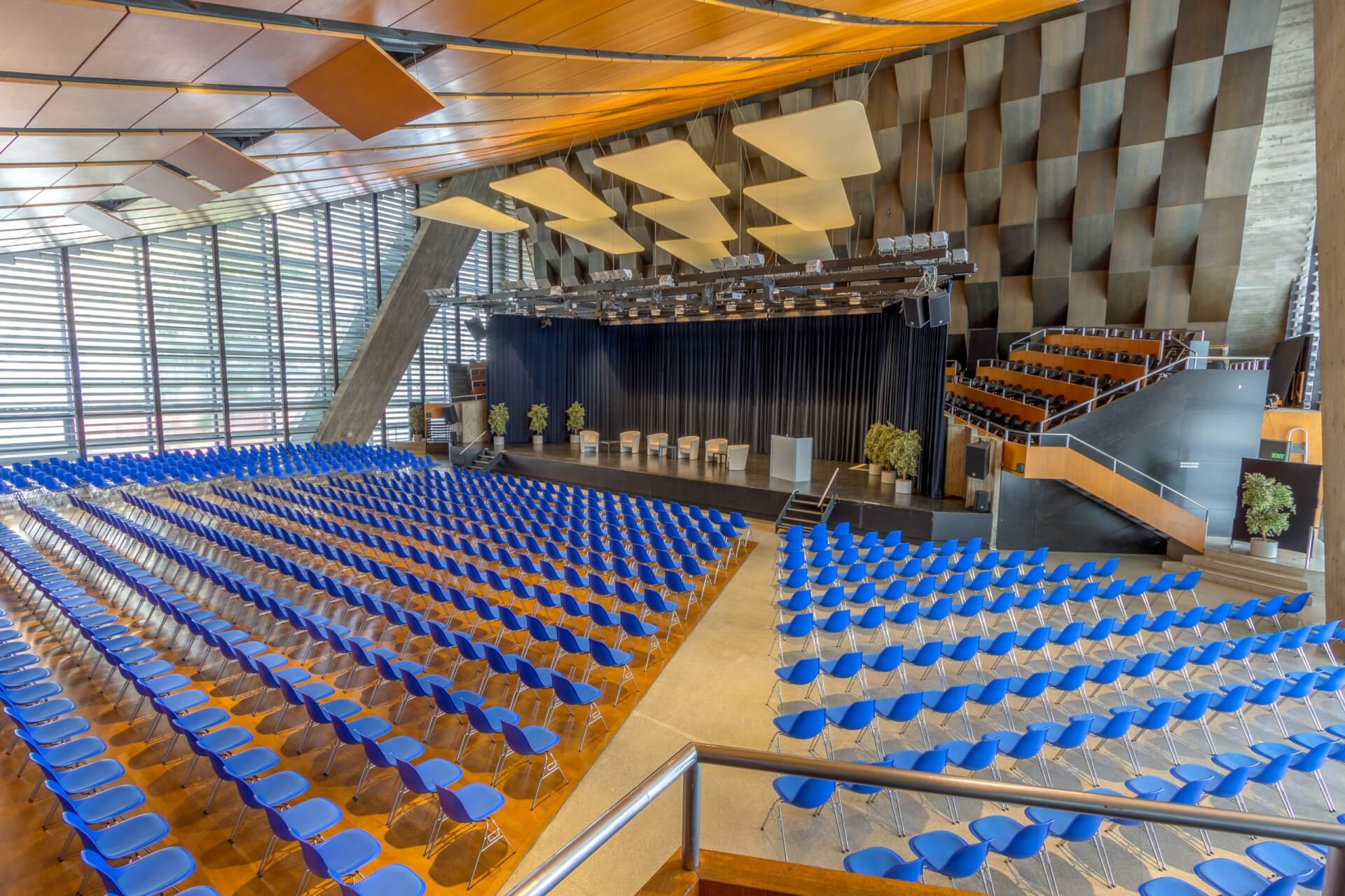 Großes Auditorium mit blauen Stuhlreihen vor einer Bühne, modernen geometrischen Wänden und abgestuften Sitzreihen auf der rechten Seite.