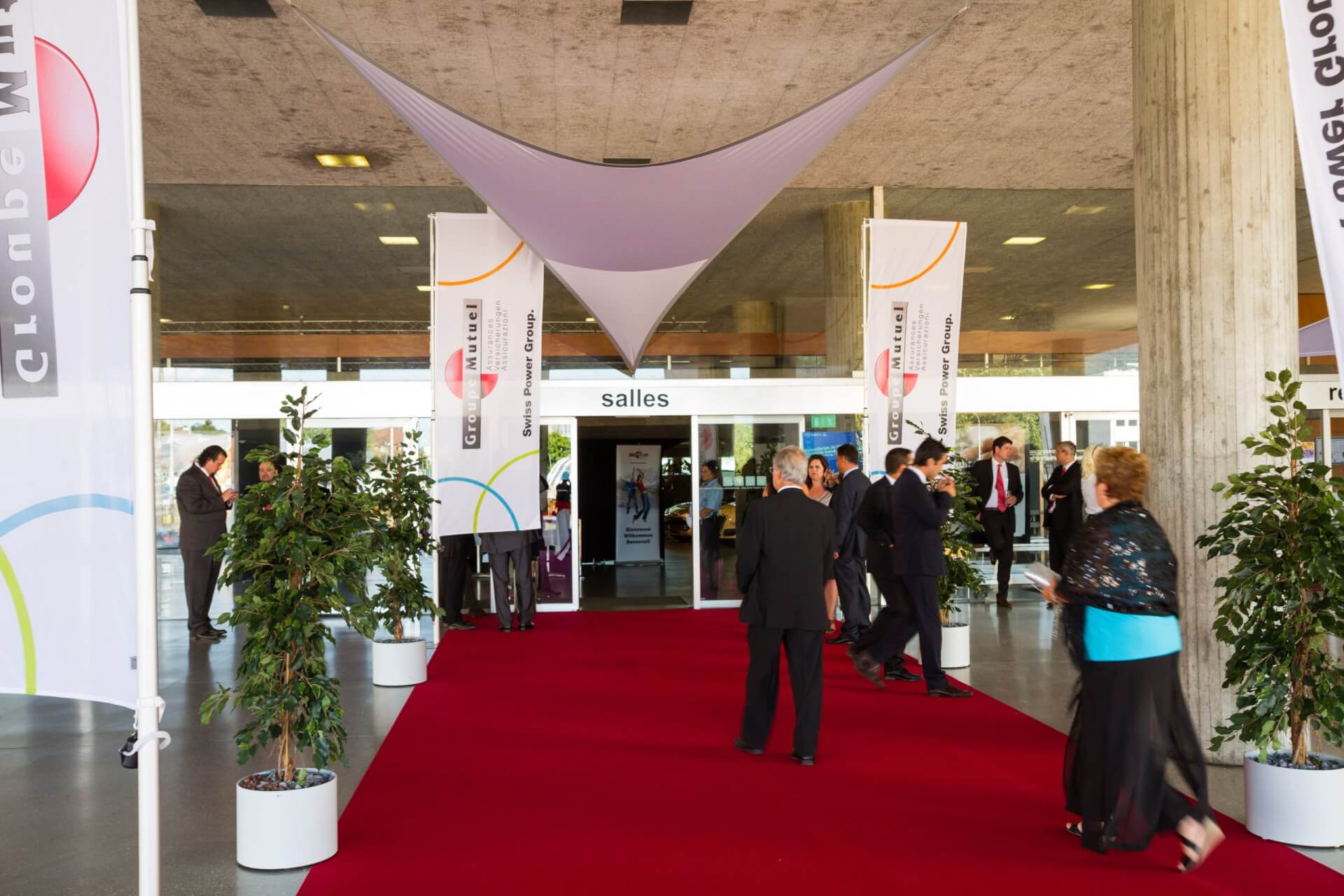 Menschen in förmlicher Kleidung gehen über einen roten Teppich, der zu Glastüren mit der Aufschrift 