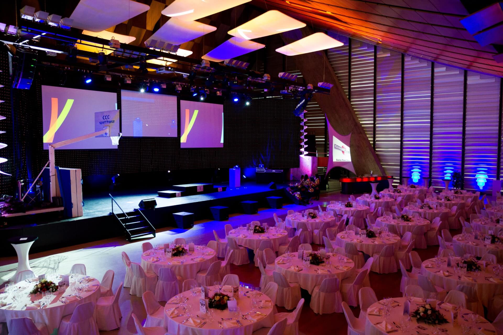Großer Bankettsaal, der für eine Veranstaltung mit runden Tischen mit weißen Tischdecken, Blumenschmuck, einer Bühne, großen Bildschirmen und violetter Hintergrundbeleuchtung eingerichtet ist.