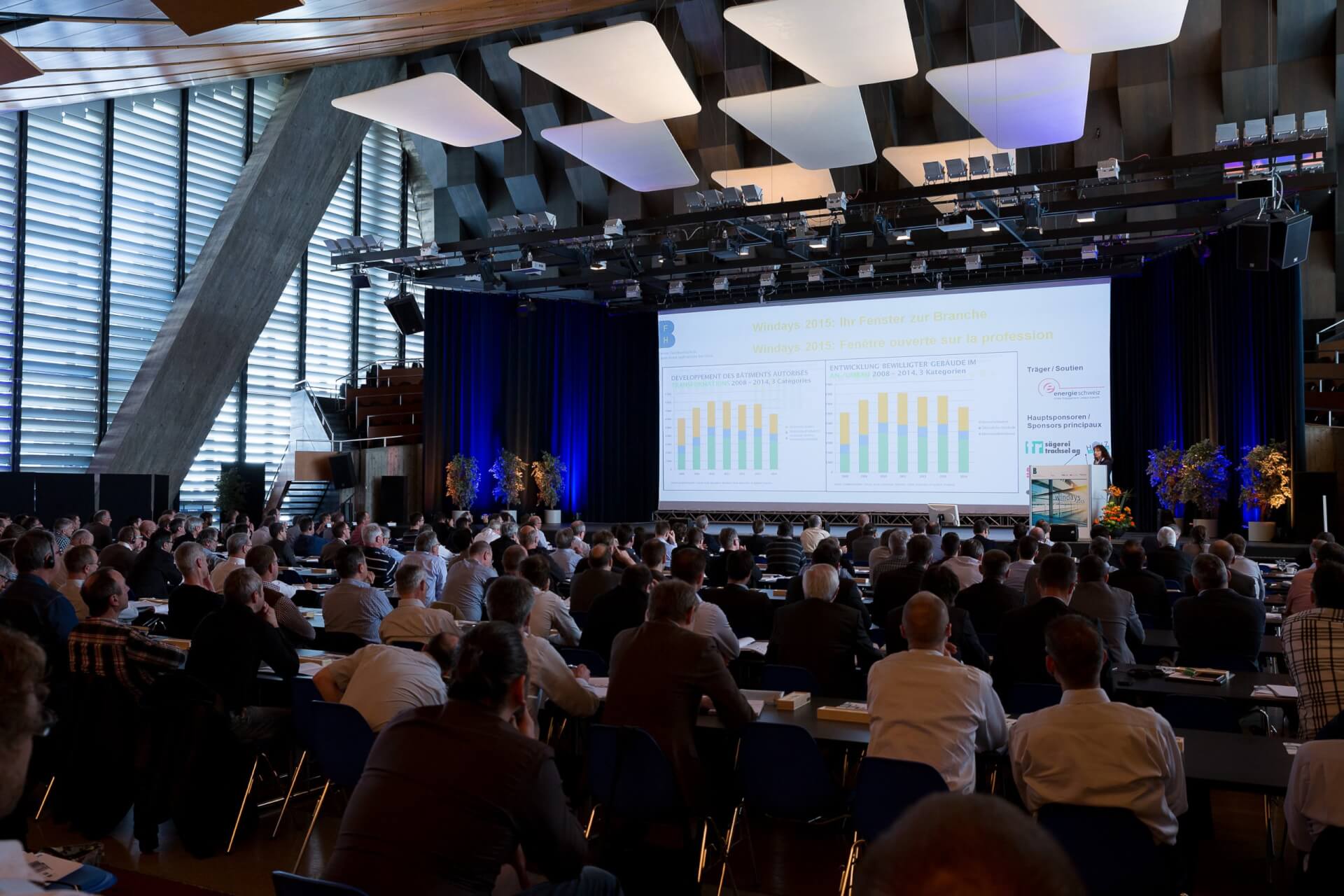 Ein großes Publikum sitzt in einem modernen Konferenzsaal und verfolgt eine Präsentation mit Diagrammen, die auf eine Leinwand im vorderen Teil des Raums projiziert werden.