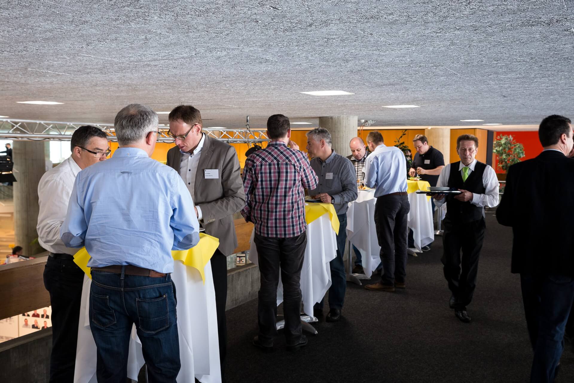 Menschen stehen und unterhalten sich um hohe Tische, die mit weißen und gelben Tischdecken bedeckt sind, bei einer Indoor-Veranstaltung; im Hintergrund trägt ein Kellner ein Tablett.