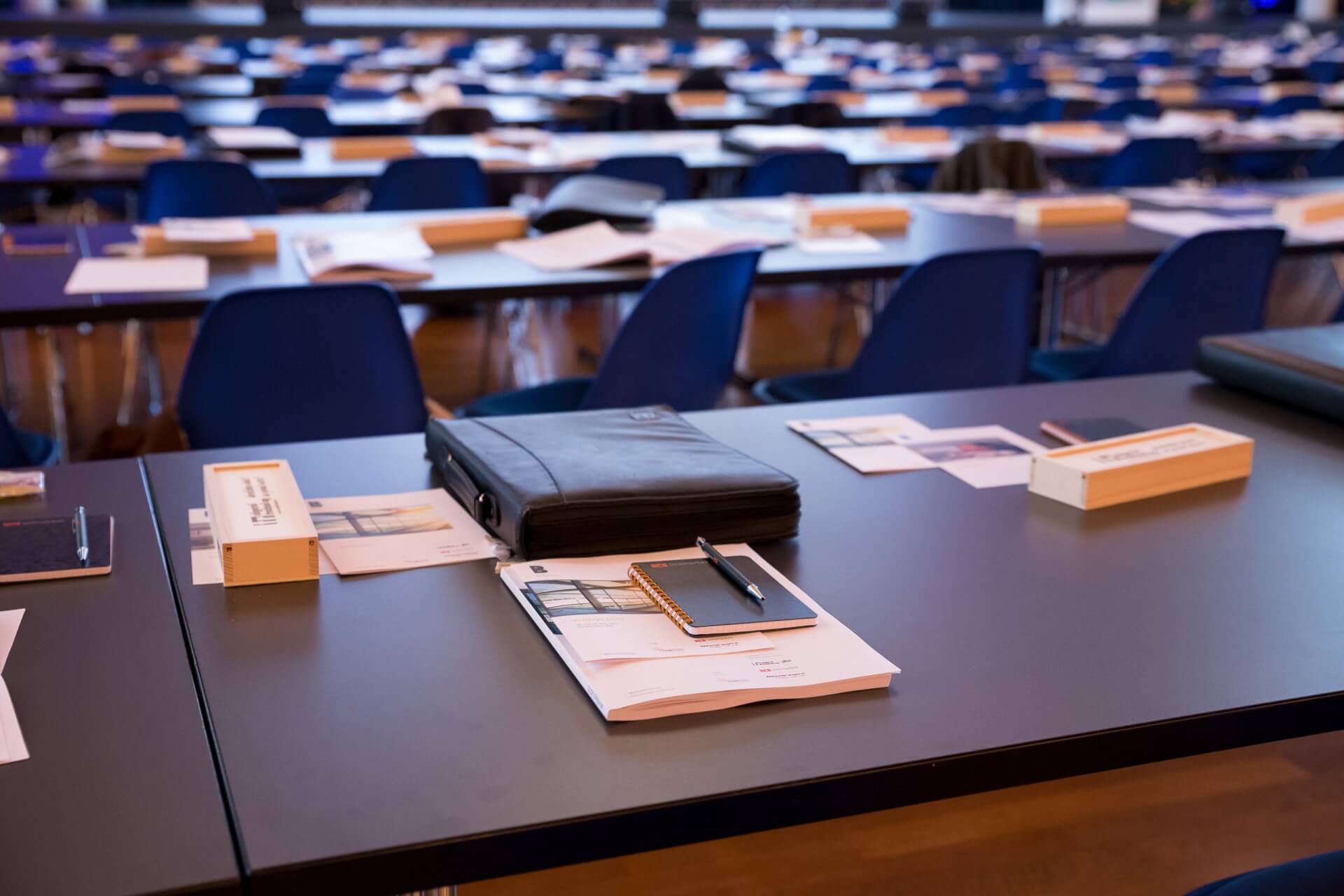 Reihen von Tischen und blauen Stühlen, die für eine Veranstaltung oder Prüfung aufgestellt sind, mit Papieren, Ordnern und Stiften, die ordentlich auf jedem Schreibtisch liegen.