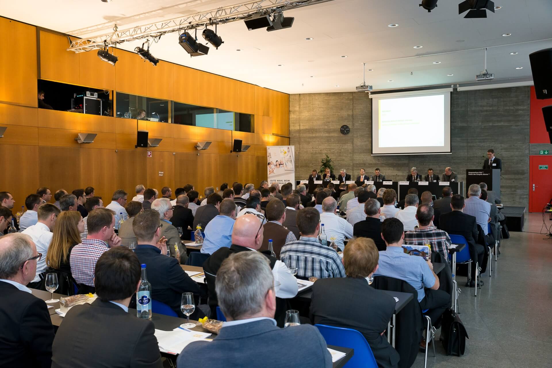 Eine große Gruppe von Personen sitzt vor einem Rednerpult in einem modernen Konferenzraum, in dem eine Präsentation an die Wand projiziert wird.