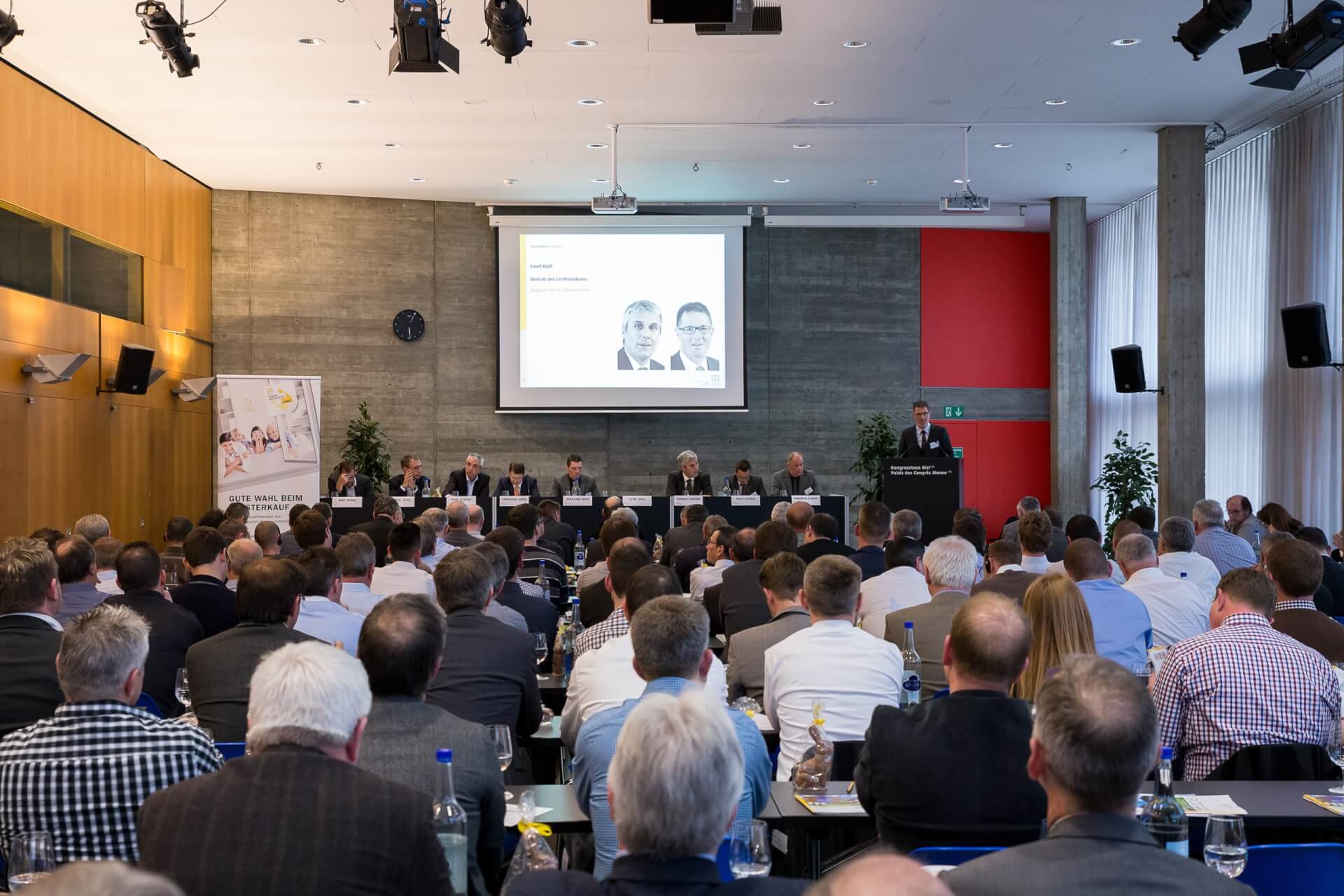 Eine große Gruppe von Personen nimmt an einer Geschäftskonferenz in einem modernen Raum teil und steht vor einer Bühne mit Rednern und einer auf eine Leinwand projizierten Präsentation.