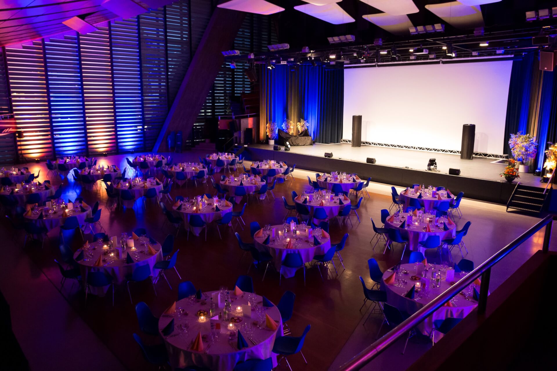 Großer Veranstaltungssaal mit runden Tischen, die für das Abendessen gedeckt sind, gedämpfter Beleuchtung mit lila und gelben Akzenten und einer Bühne mit einer großen weißen Leinwand an der Stirnseite.
