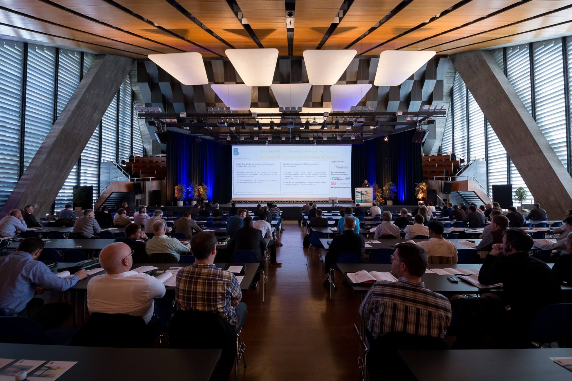 Eine große Gruppe von Personen sitzt in einem modernen Konferenzsaal vor einer Bühne, auf der eine Präsentation auf eine Leinwand projiziert wird.