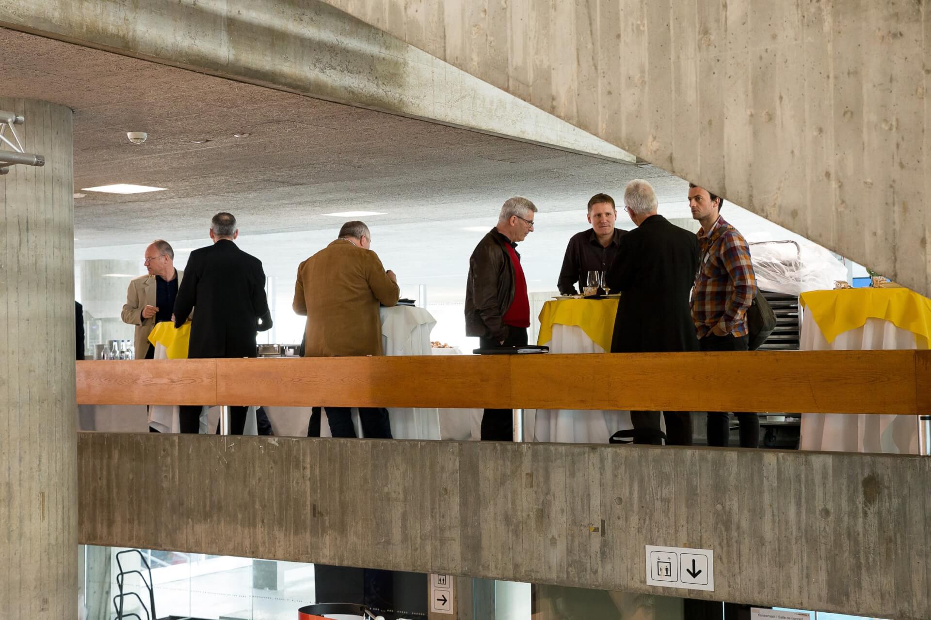 Mehrere Männer stehen um hohe, mit gelben und weißen Tischtüchern bedeckte Tische in einem Korridor eines Betongebäudes und unterhalten sich.