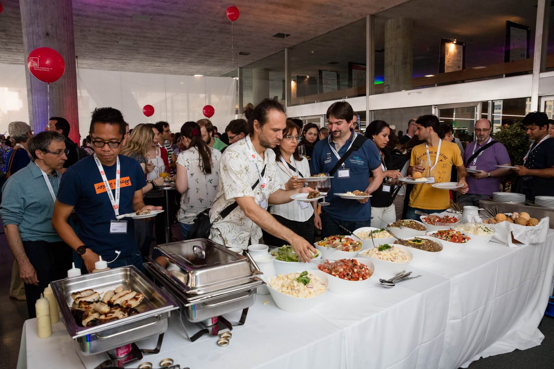 Eine Gruppe von Personen bedient sich bei einer Indoor-Veranstaltung an einem Buffet, an dem verschiedene Gerichte und Salate angeboten werden.