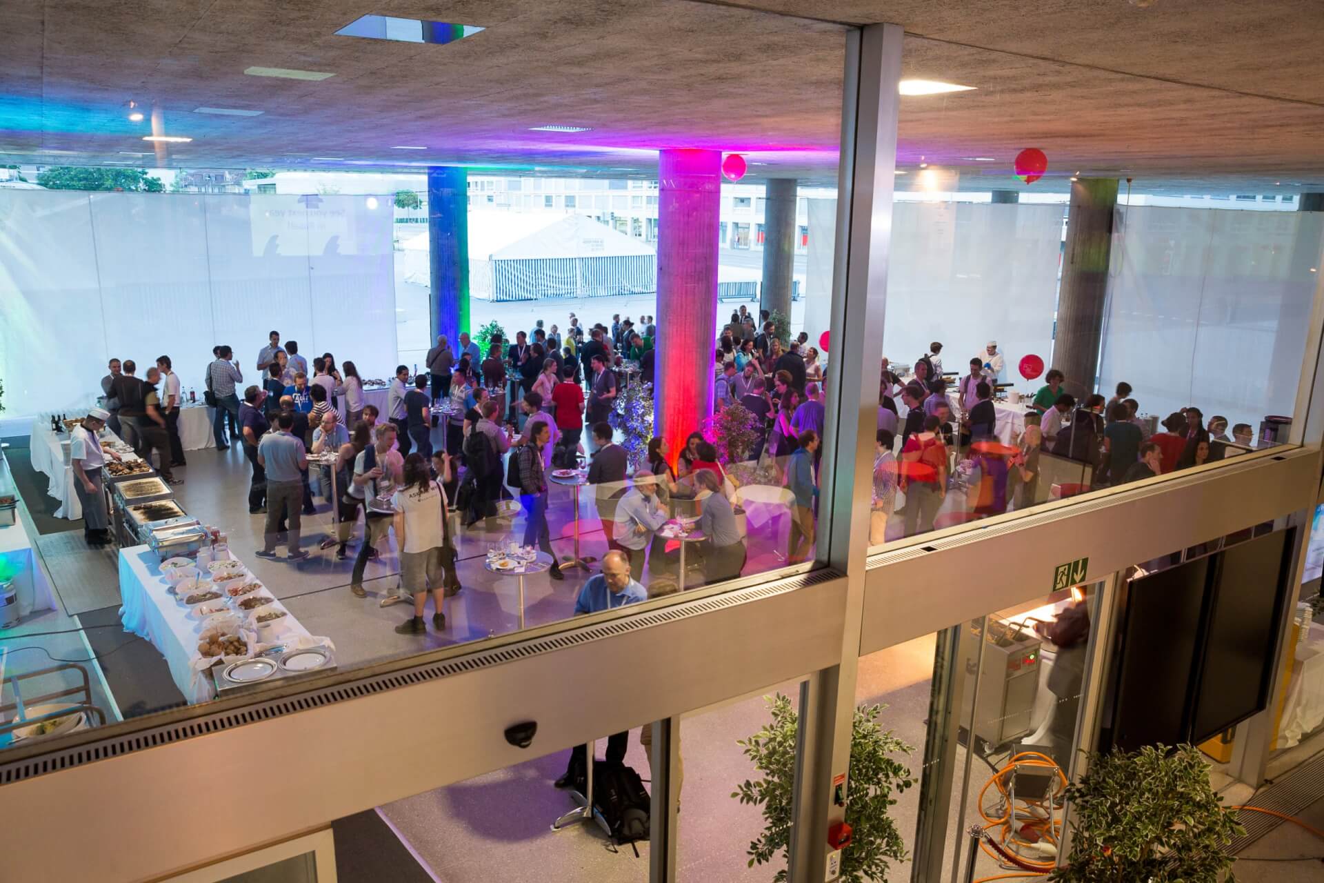 Eine große Gruppe von Menschen versammelt sich in einem modernen, überdachten Veranstaltungsort mit Tischen, die mit lila und blauer Beleuchtung ausgestattet sind und durch Glasscheiben betrachtet werden können.