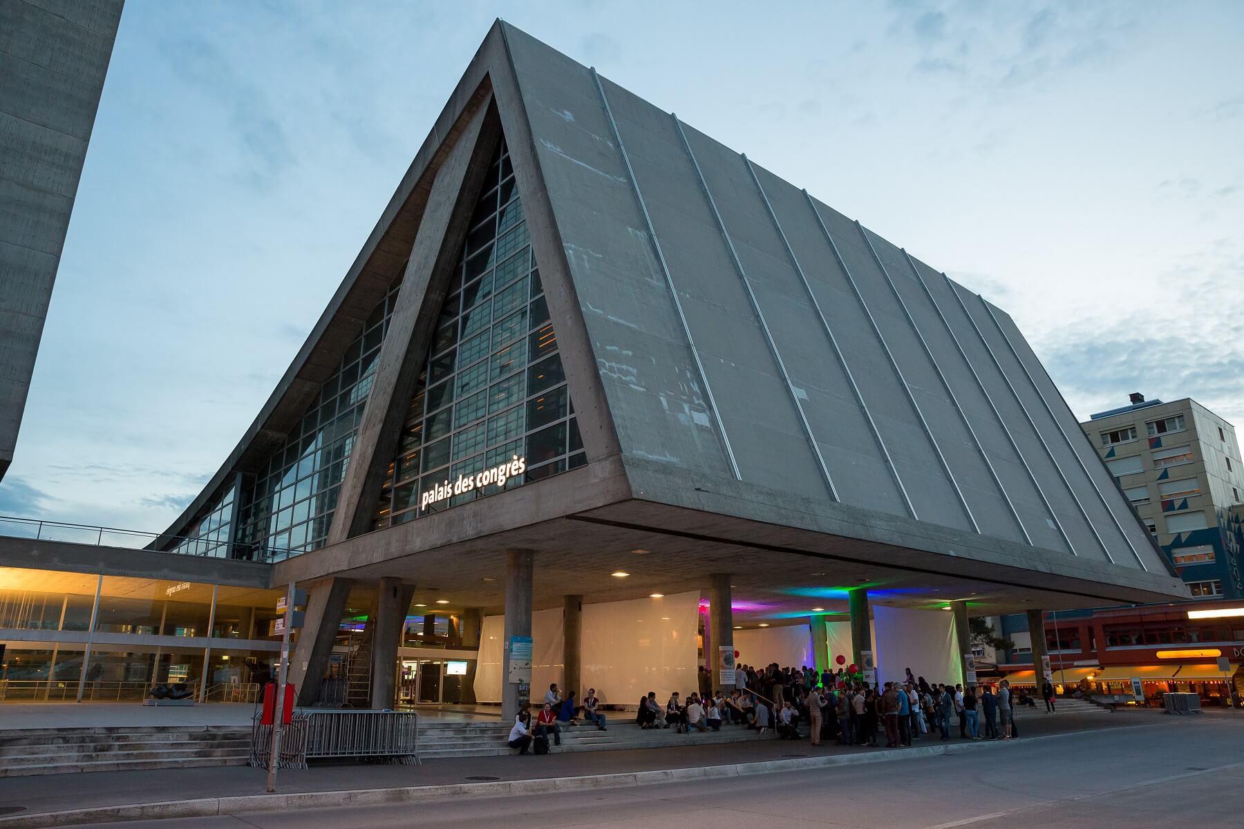 Ein modernes, kantiges Betongebäude mit großen Glasfenstern trägt die Aufschrift palais des congrès, unter dessen überdachtem Eingang sich in der Abenddämmerung Menschen versammeln.