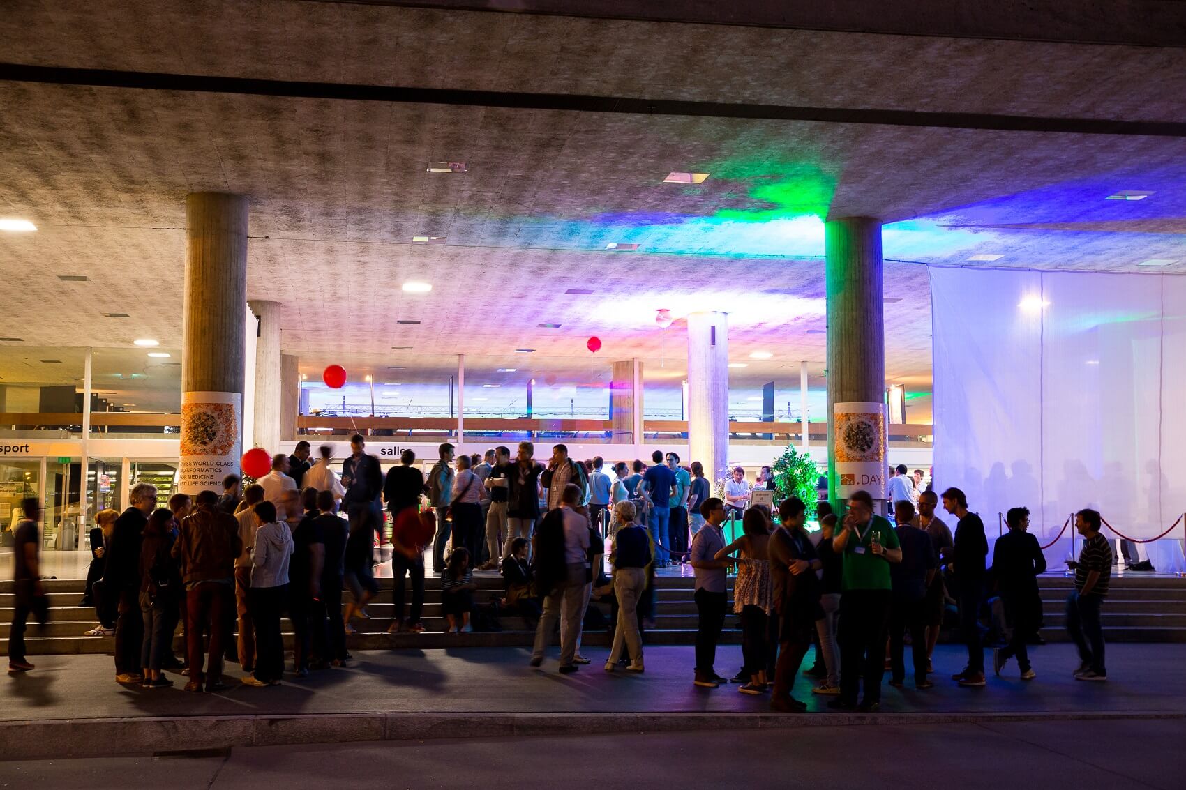 Eine große Gruppe von Menschen versammelt sich nachts vor einem modernen Gebäude unter einem Betonüberhang, der mit bunten Lichtern beleuchtet ist.