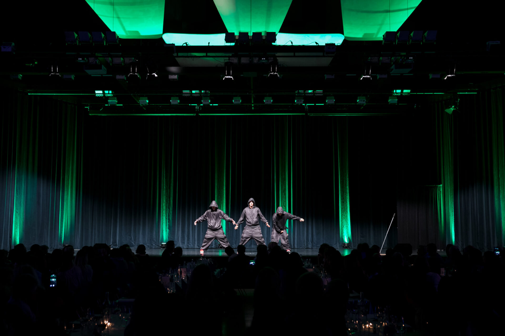 Drei Tänzer treten auf einer grün beleuchteten Bühne auf, während das Publikum von Tischen im Vordergrund aus zusieht.