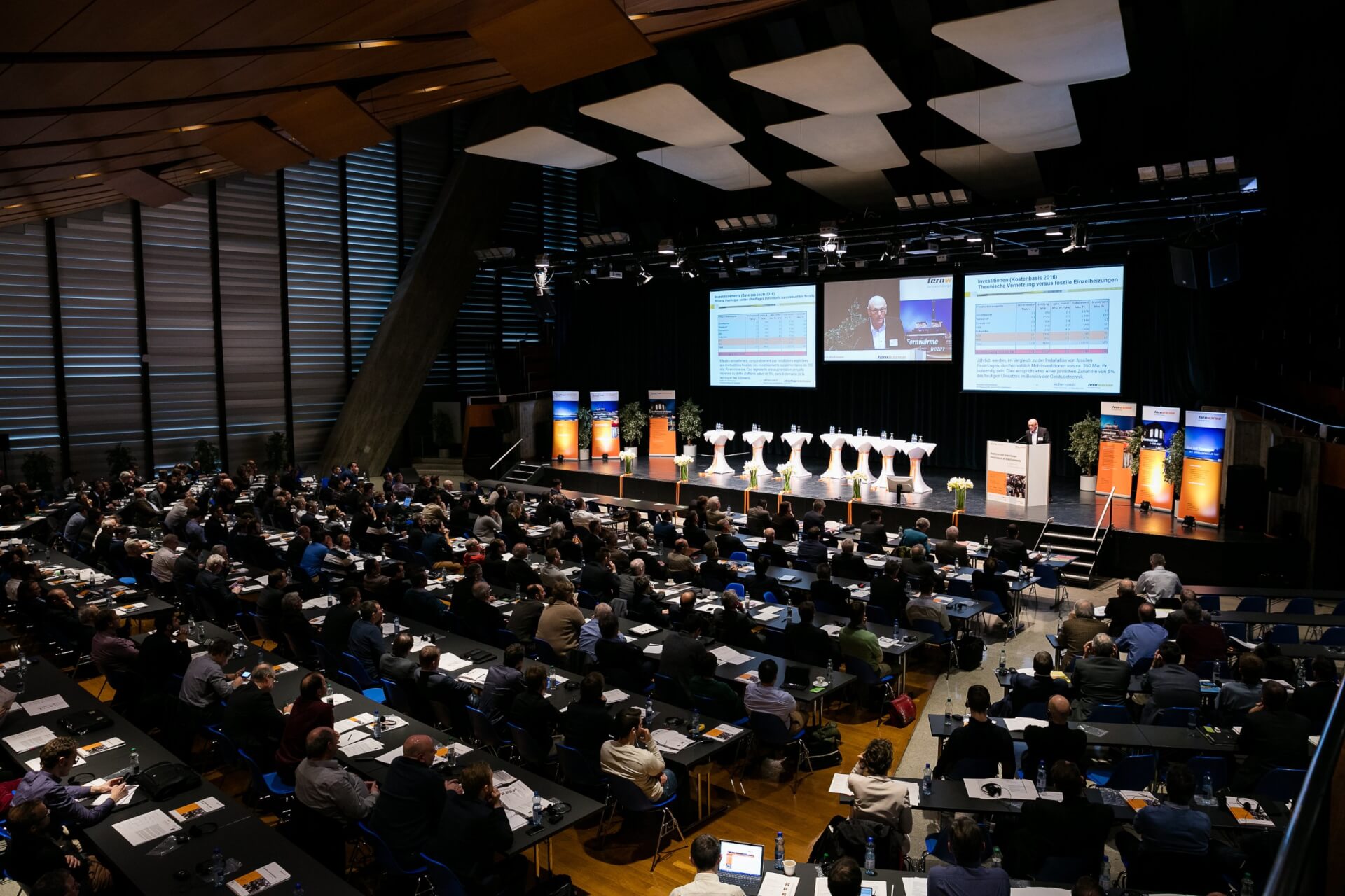 Ein großer Konferenzraum mit sitzenden Teilnehmern, die einer Bühne mit einem Redner, Diskussionsteilnehmern und Präsentationsfolien auf großen Bildschirmen gegenüberstehen.