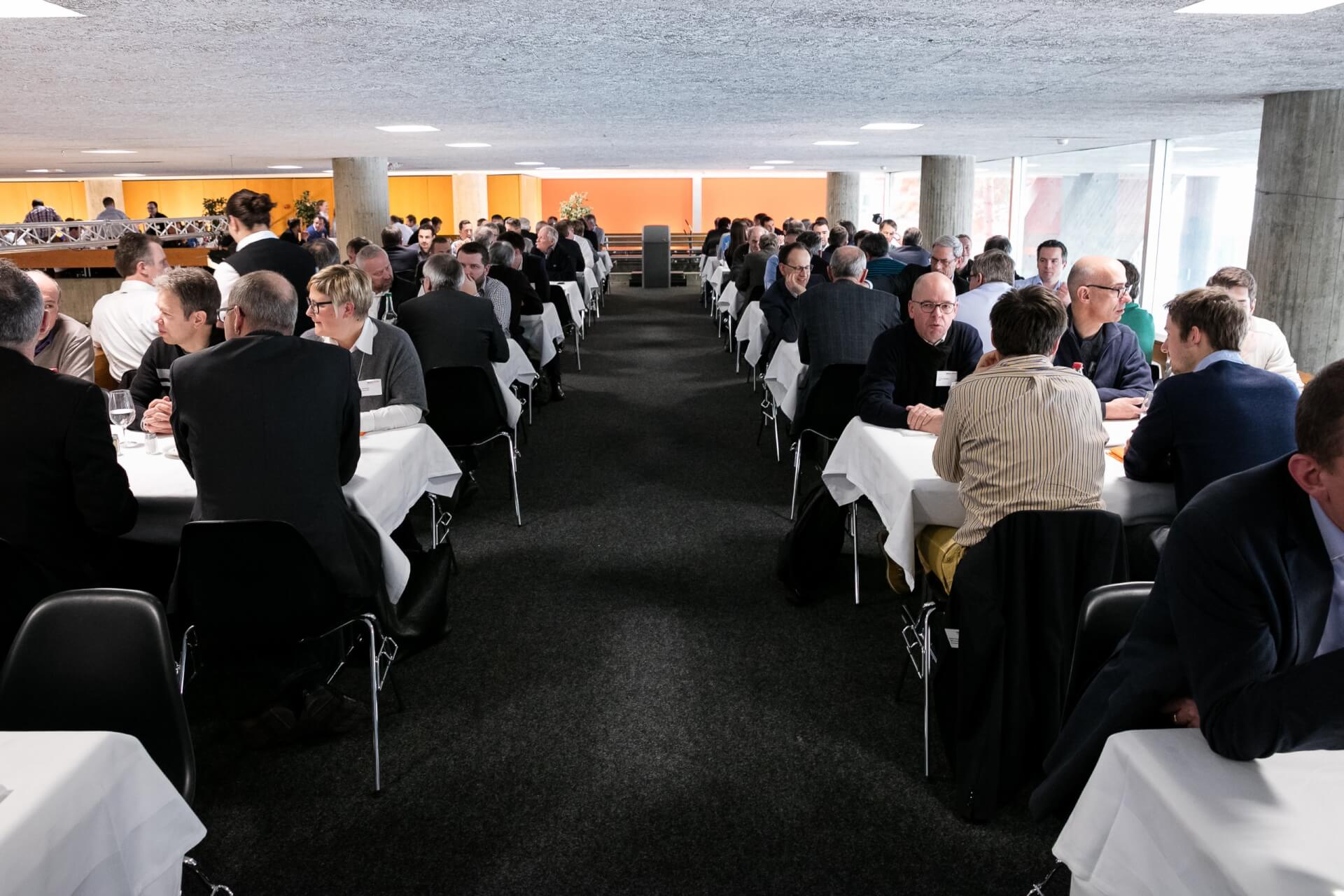 Menschen, die in Reihen an langen Tischen in einem Konferenz- oder Geschäftssitzungsraum sitzen und sich unterhalten.