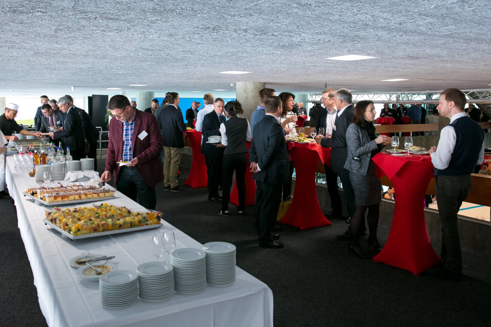 Menschen stehen um rote Cocktailtische herum und unterhalten sich bei einer Buffetveranstaltung, wobei verschiedene Essenstabletts, Getränke und Teller auf einem langen Tisch im Vordergrund zu sehen sind.