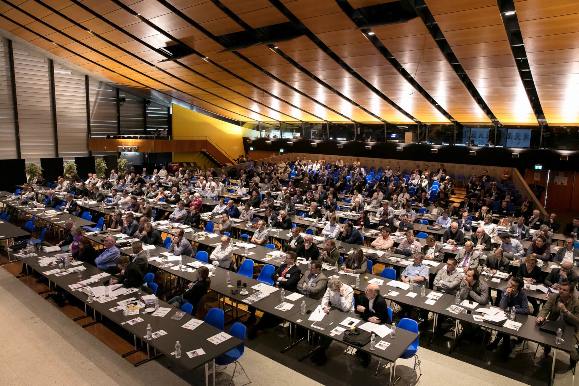 Eine große Gruppe von Personen sitzt an Tischen in einem modernen Konferenzsaal, mit Blick nach vorne, und auf den Tischen liegen Papiere und Laptops.