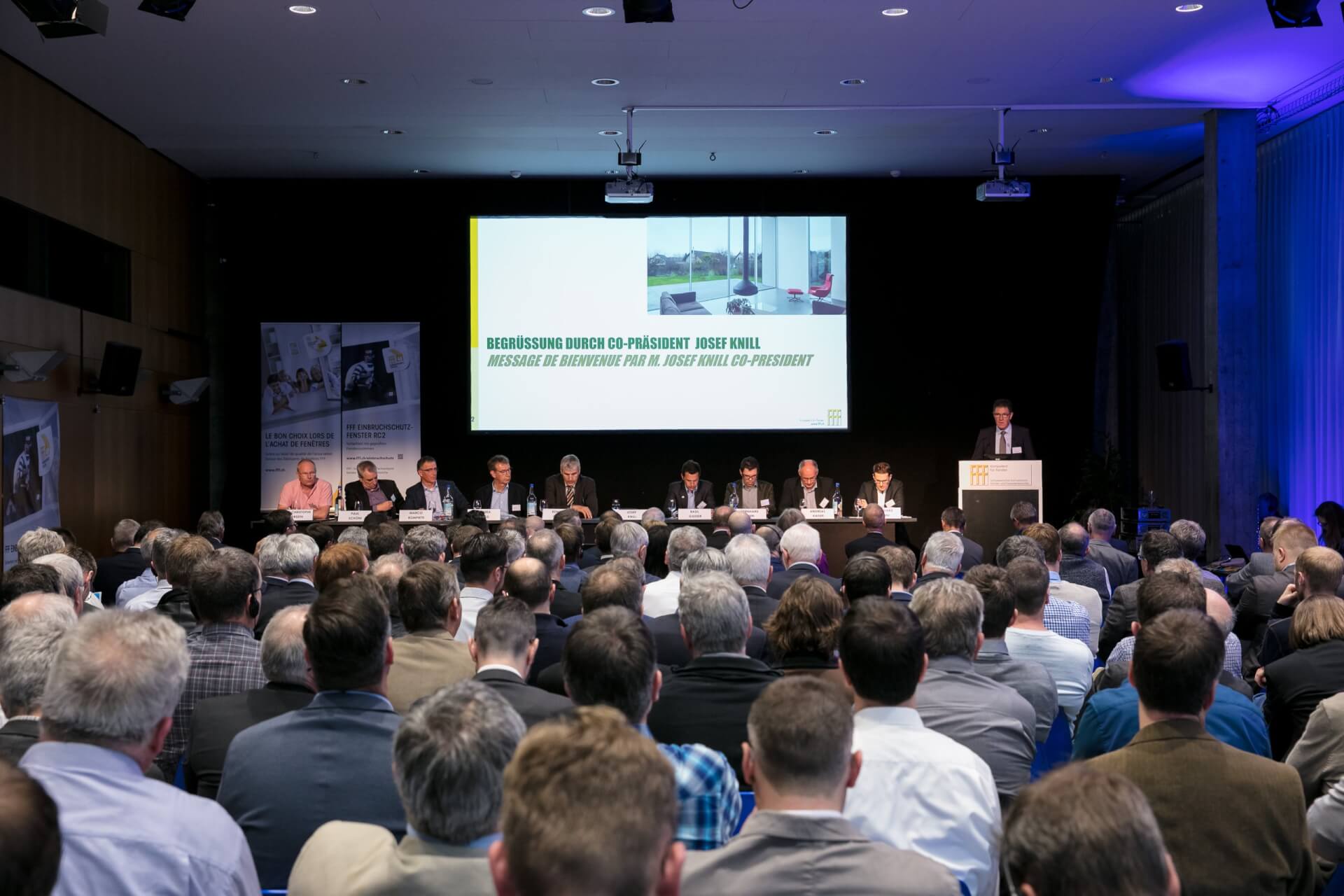 Ein großes Publikum sitzt vor einer Bühne mit einer Gruppe von Rednern und einem Moderator auf einem Podium, während auf einem Bildschirm eine Begrüßungsbotschaft angezeigt wird.