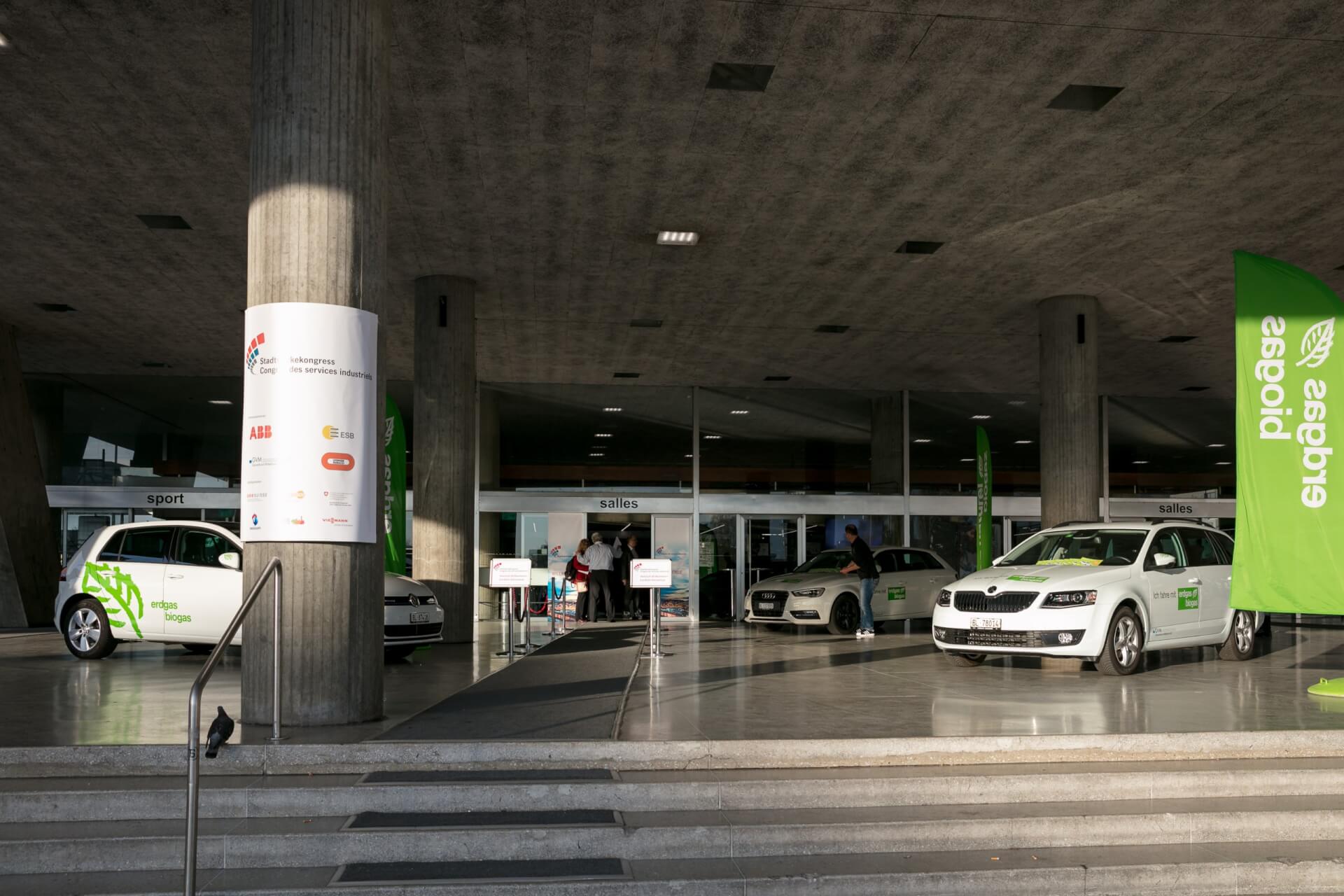 Drei weiße Elektroautos sind auf einer Betonrampe vor einem Gebäudeeingang geparkt und mit grünen Flaggen und Schildern gekennzeichnet.