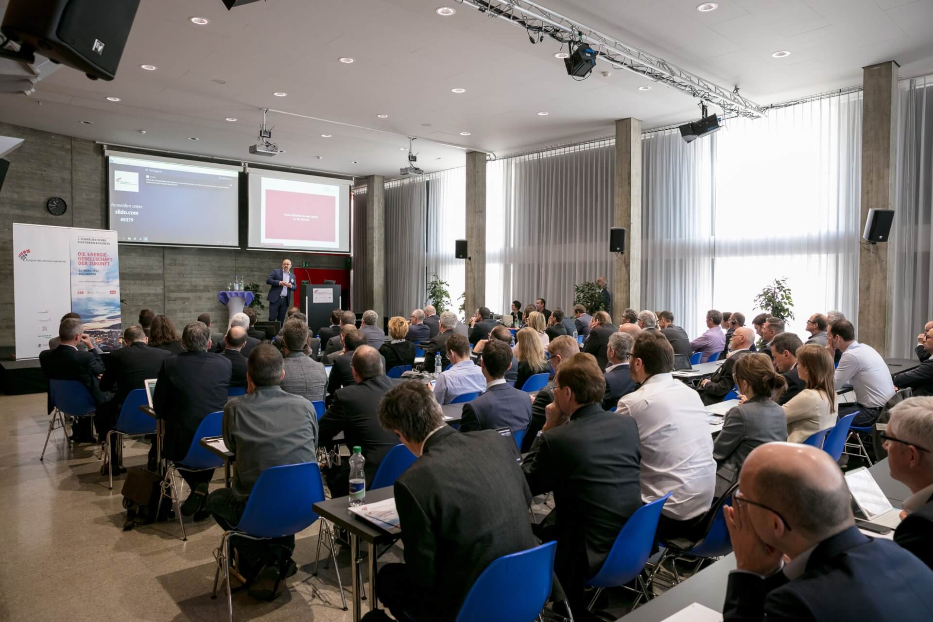 Eine Gruppe von Personen sitzt in einem Konferenzraum und hört einem Redner zu, der im vorderen Teil des Raums Folien präsentiert.