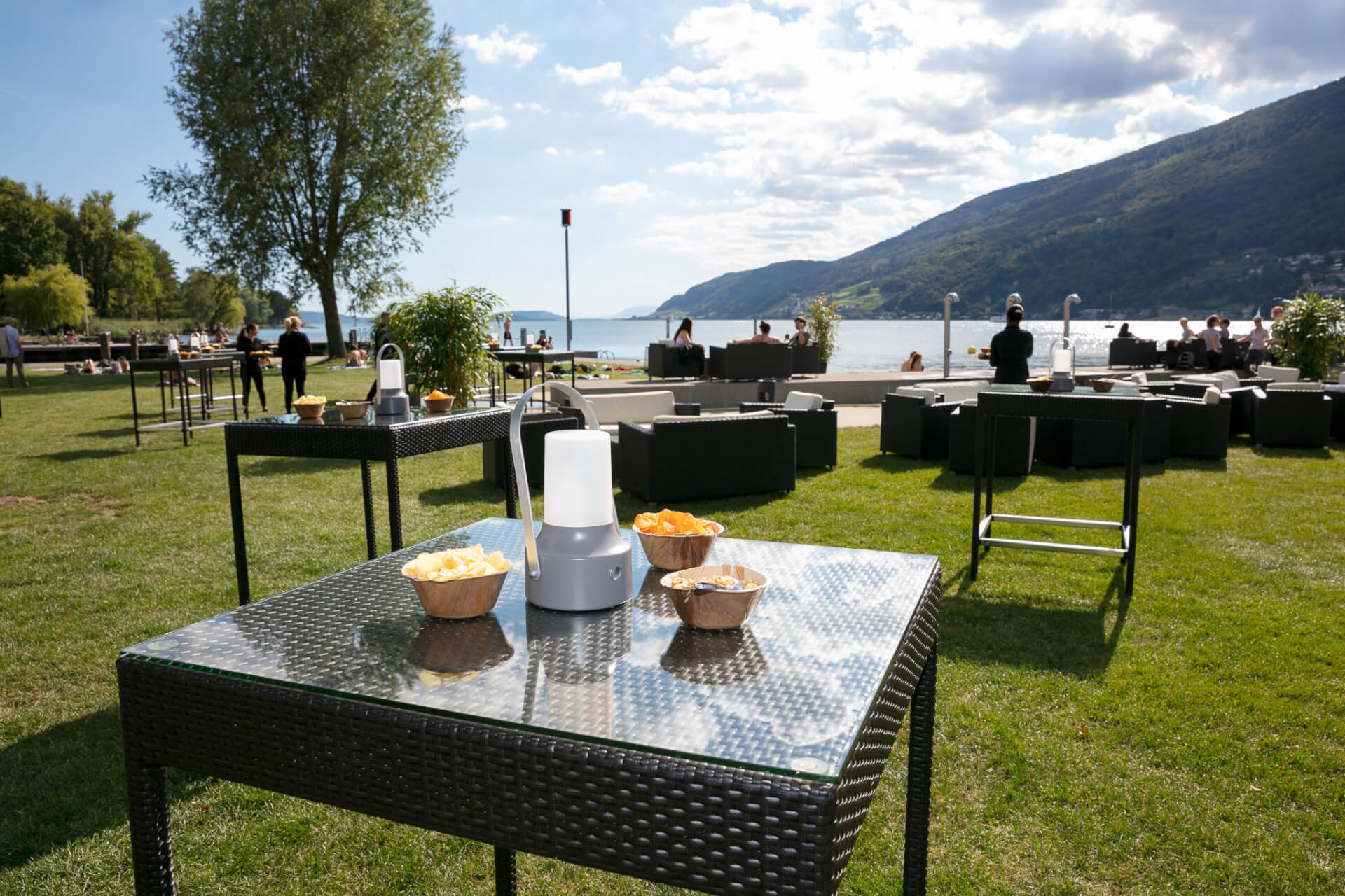 Sièges extérieurs avec tables en osier et snacks sur une pelouse au bord d'un lac, avec des montagnes et des gens en arrière-plan sous un ciel partiellement nuageux.