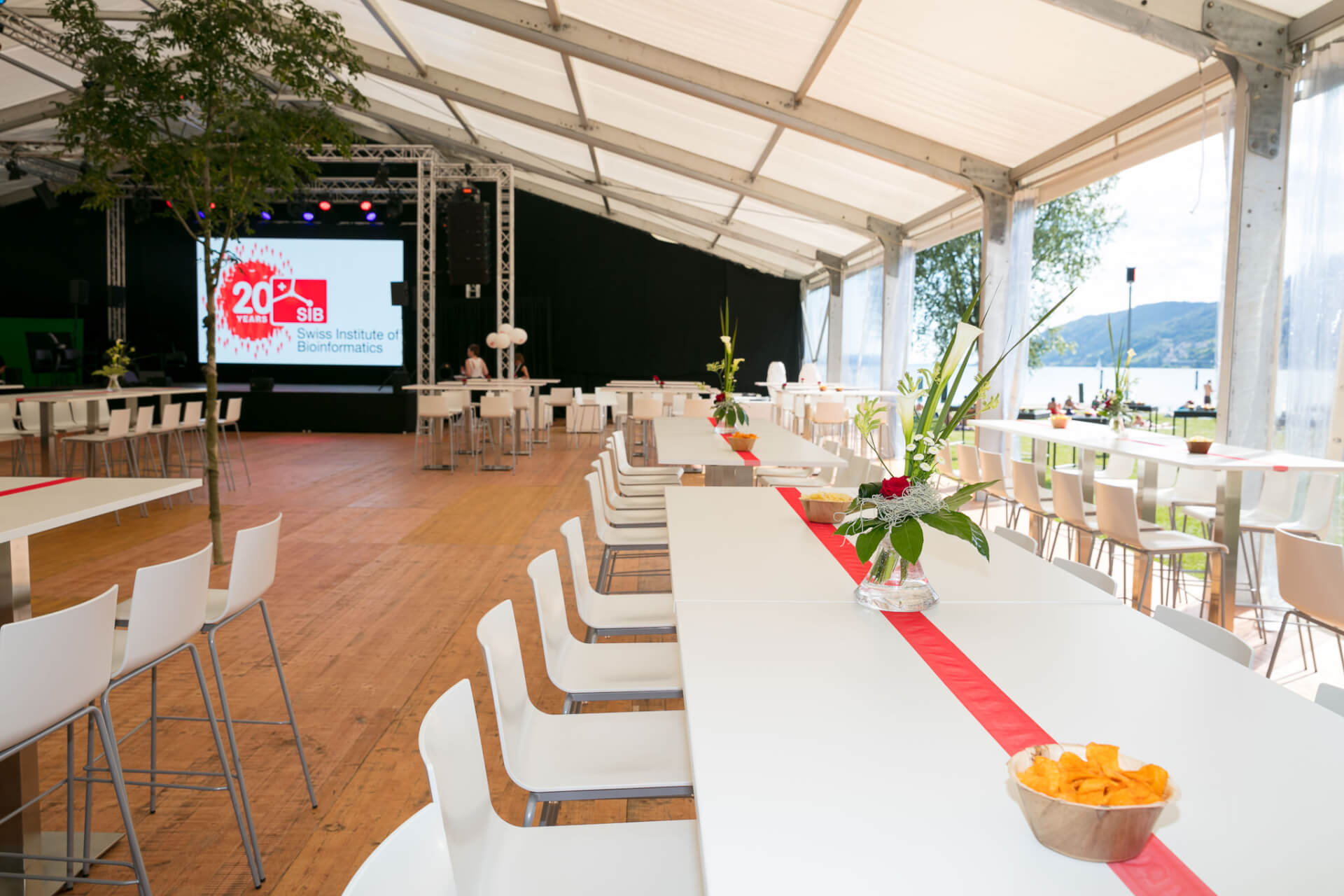 Ein grosses Veranstaltungszelt mit weissen Tischen und Stühlen, schlichtem Blumenschmuck, einer Snackschale und einer Bühne mit einer Leinwand, auf der das 20. SIB Schweizerisches Institut für Bioinformatik zu sehen ist.