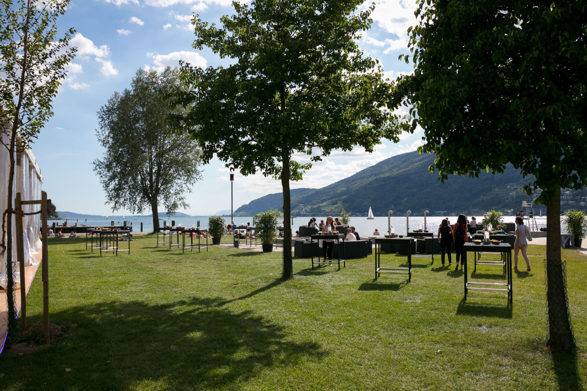 Die Menschen versammeln sich auf einer grasbewachsenen Fläche am Seeufer mit Sitzgelegenheiten im Freien, Bäumen und fernen Bergen unter einem teilweise bewölkten Himmel.