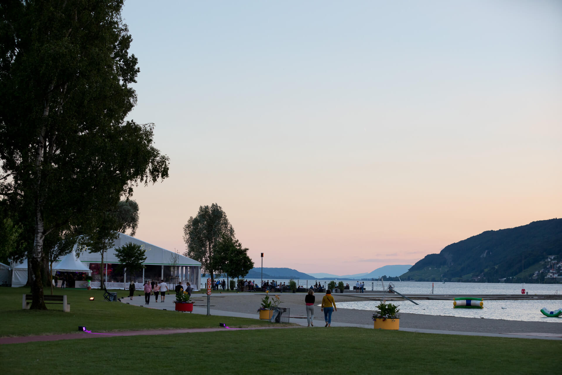 Des personnes marchent le long d'une promenade au bord d'un lac au coucher du soleil, avec des tentes, des arbres et des collines lointaines visibles sous un ciel clair.
