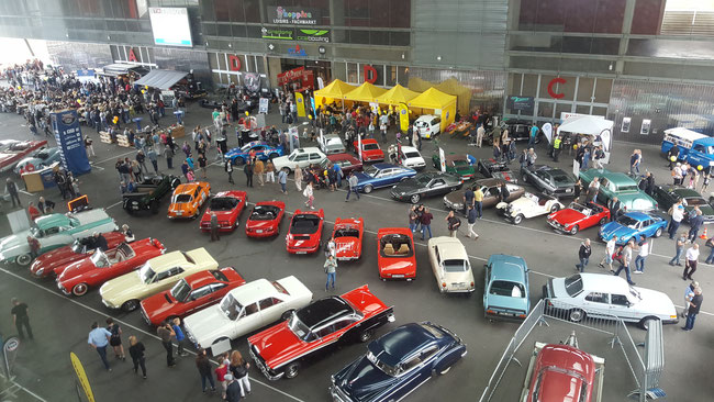 Eine große überdachte Autoshow mit zahlreichen Oldtimern, herumlaufenden Menschen, Verkaufsständen mit gelben Zelten und einer Menschenmenge, die sich am Eingang versammelt hat.
