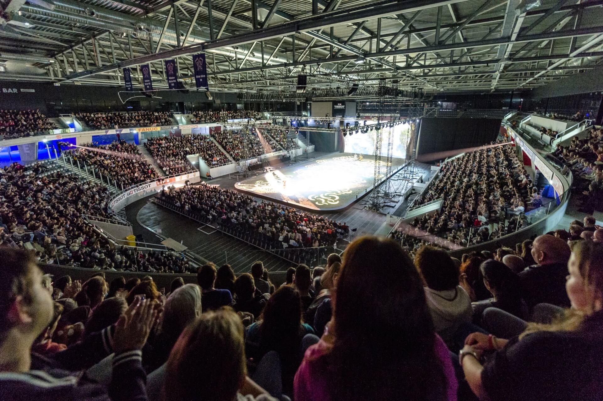 Ein großes Publikum füllt eine Halle und verfolgt eine Aufführung auf einer hell erleuchteten Bühne in der Mitte des Raums.